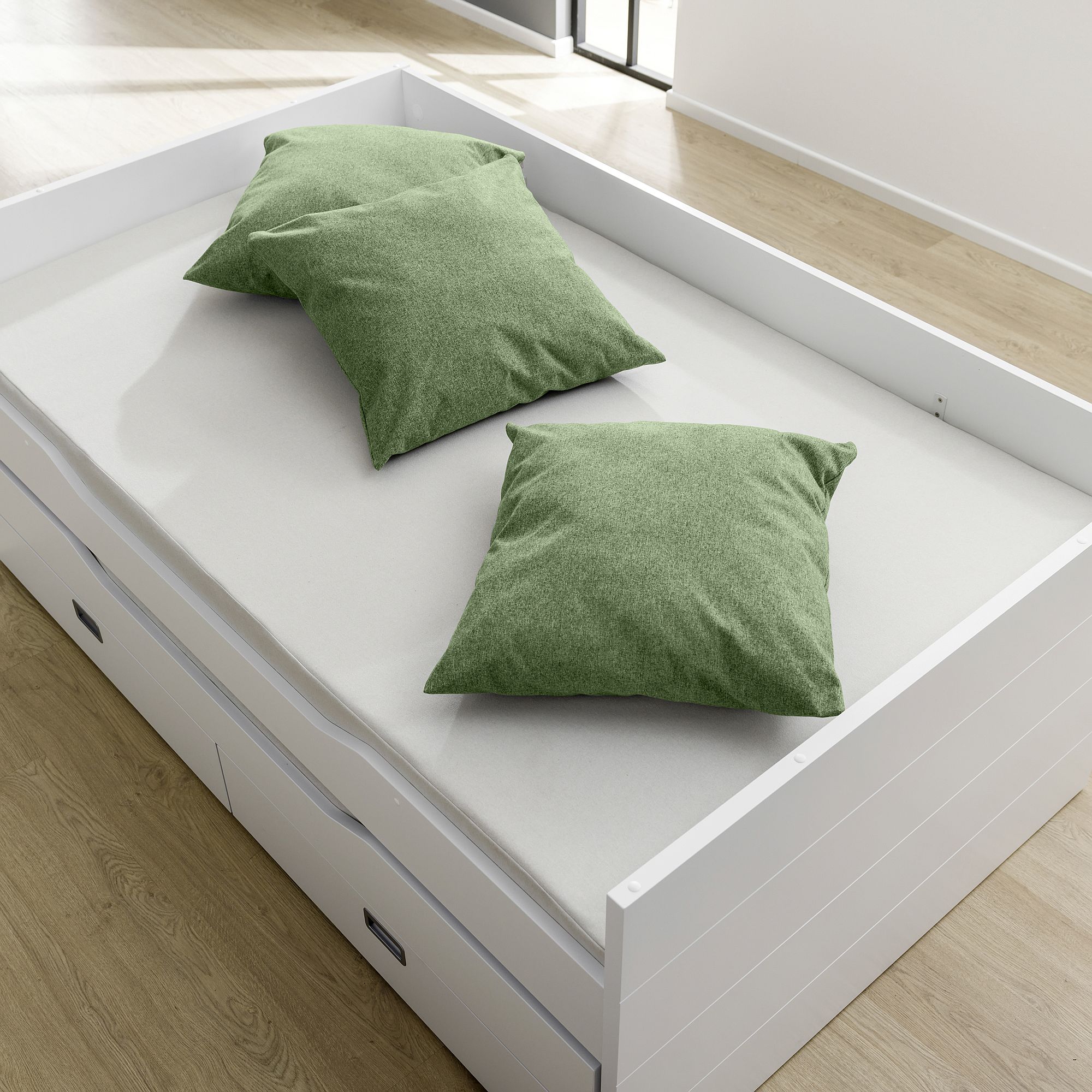 White daybed with a light mattress and two green pillows on a wooden floor.