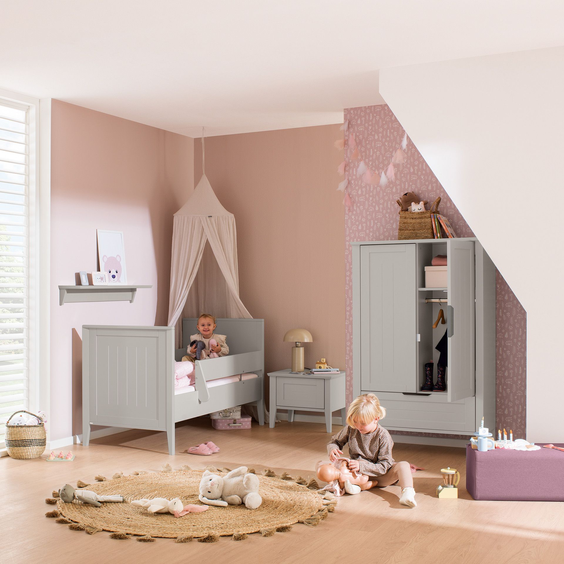 Zwei Kinder spielen in einem rosa-grauen Kinderzimmer mit Spielzeug und Möbeln.