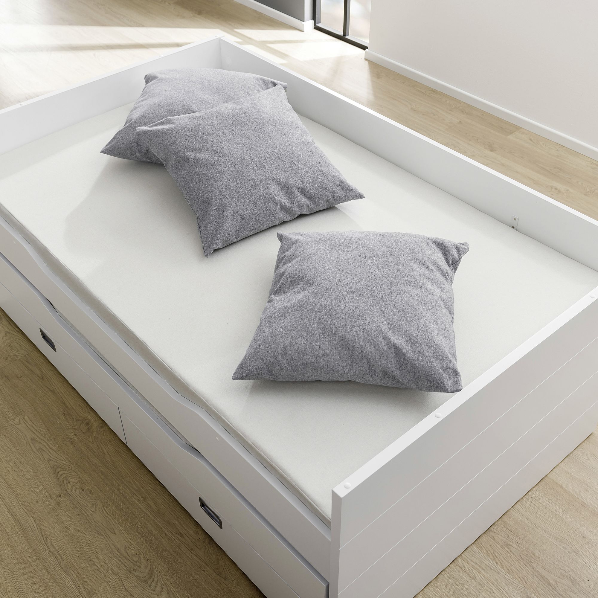 A white bed with two gray pillows on a light wood floor.