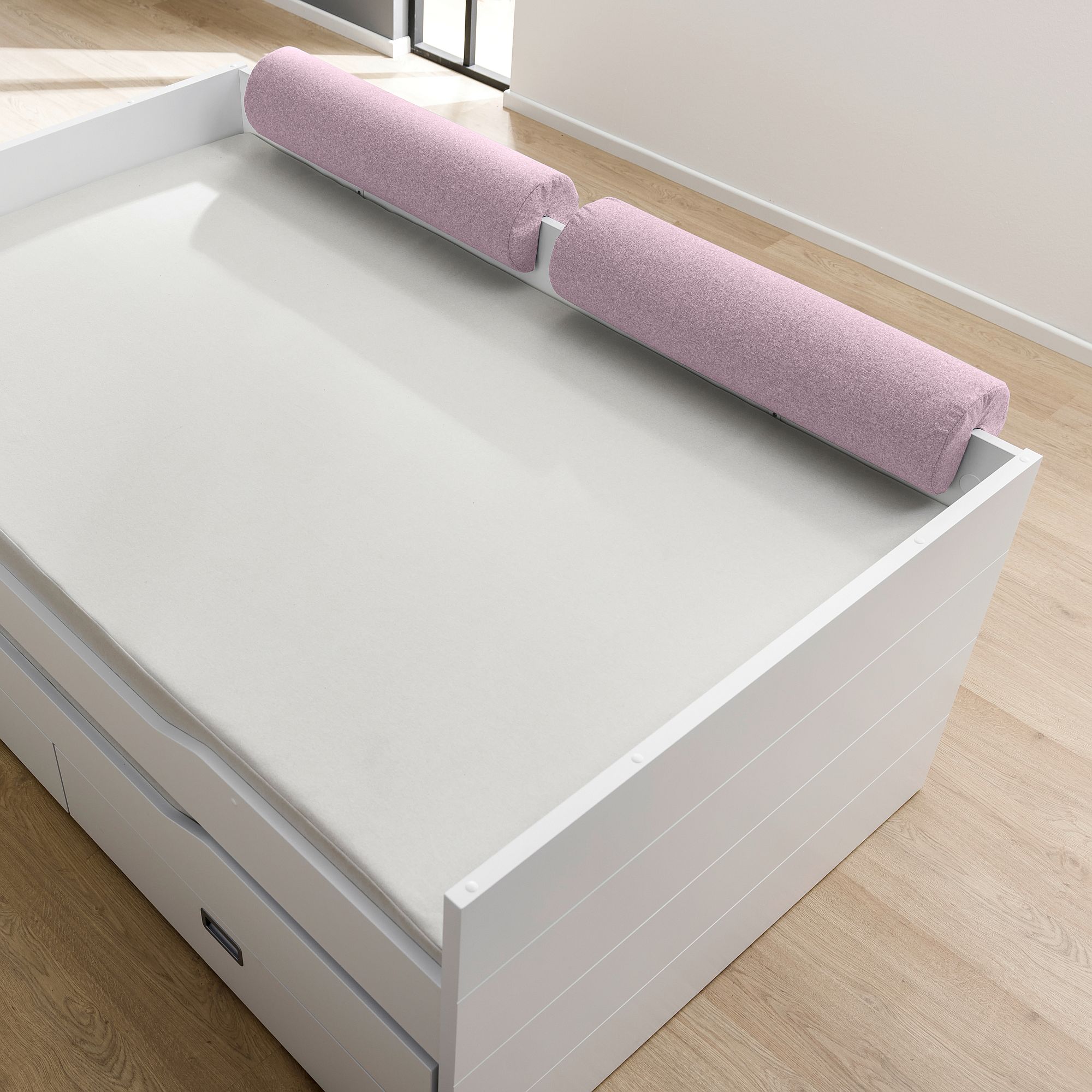 White bed with light pink bolster pillows along the back edge, on a wood floor.