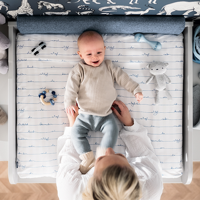 A smiling baby lies on a changing table as an adult holds their legs.
