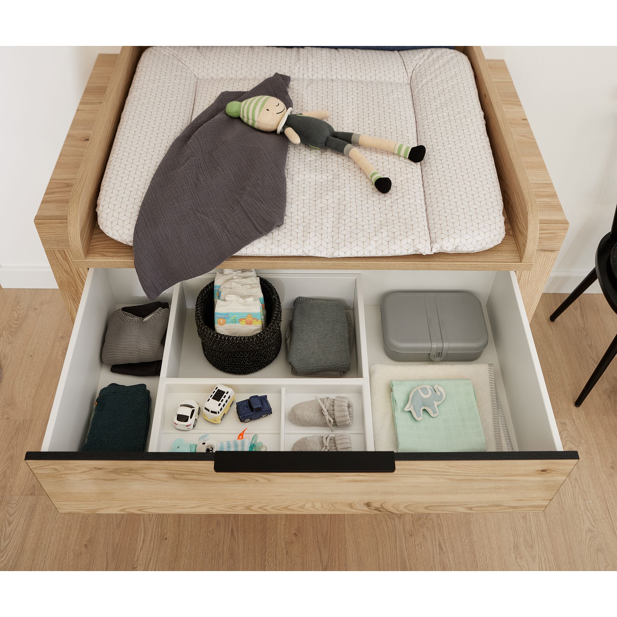 Offene Schublade unter dem Wickeltisch mit Babykleidung, Spielzeug und Windeln.