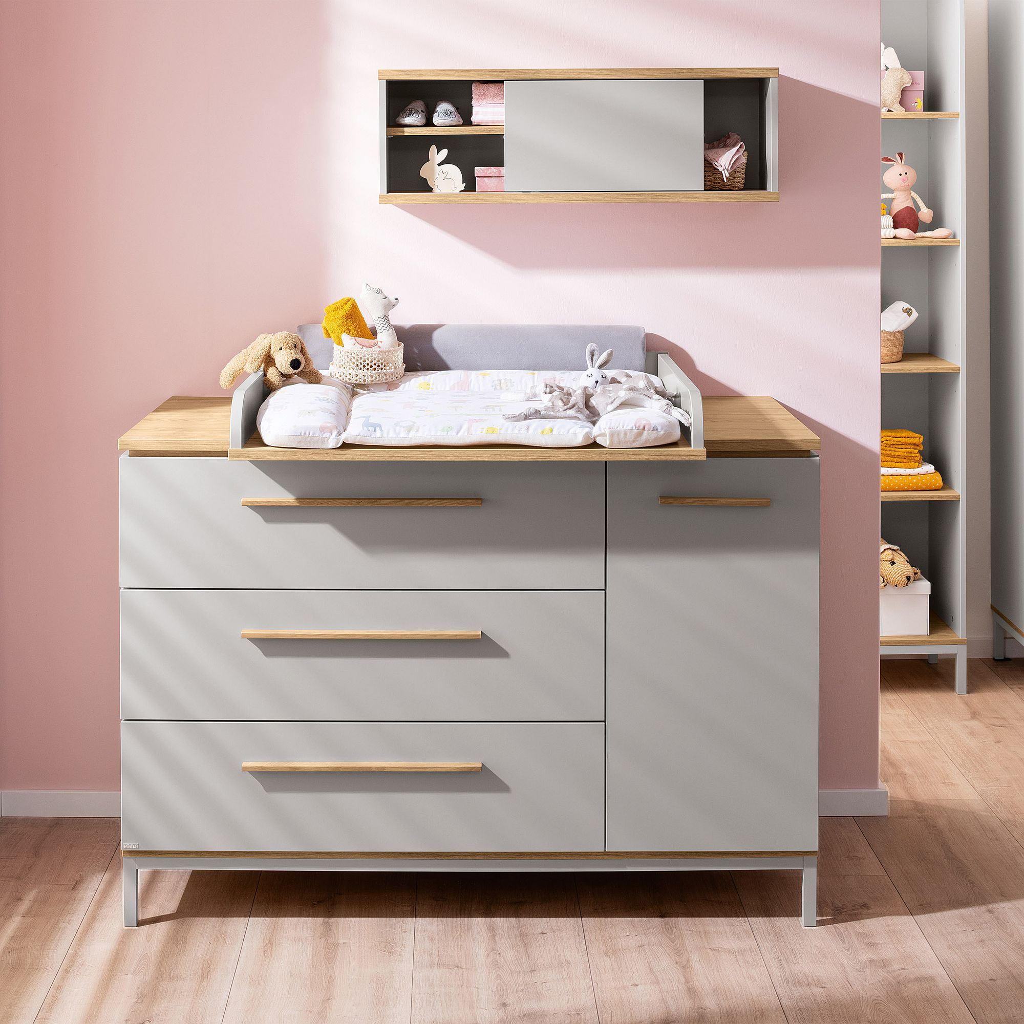 Moderner Wickeltisch mit Spielzeug und Regalen vor einer rosa Kinderzimmerwand.