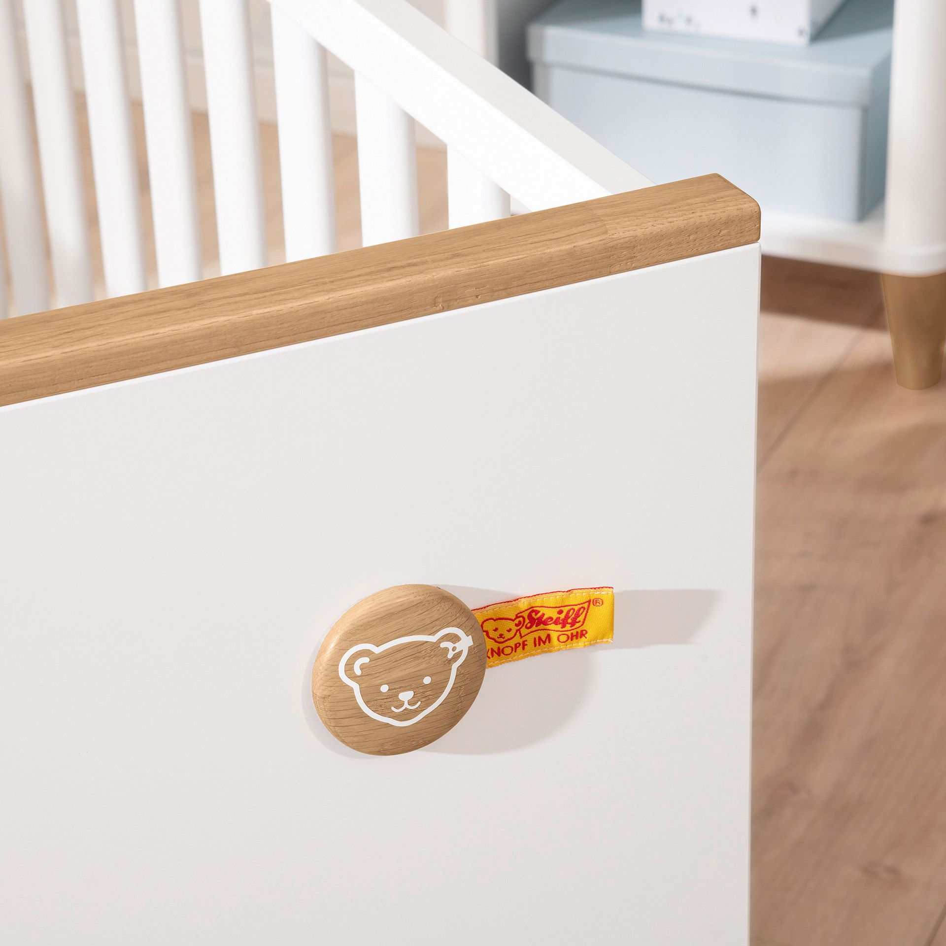 Babybett Lotte & Fynn - Steiff by PAIDI 70 x 140 cm Nahaufnahme einer weißen Krippe mit einem hölzernen Bärengesicht und einem gelben Anhänger.