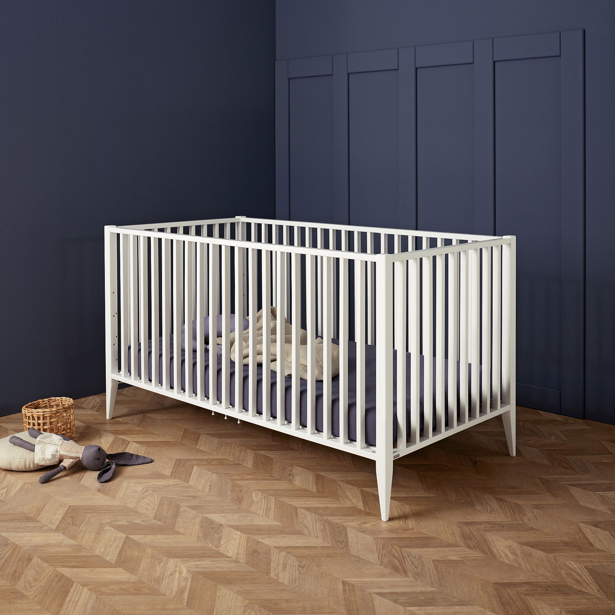 Weißes Kinderbett mit Bettzeug in einem marineblauen Zimmer mit Holzboden im Fischgrätmuster.
