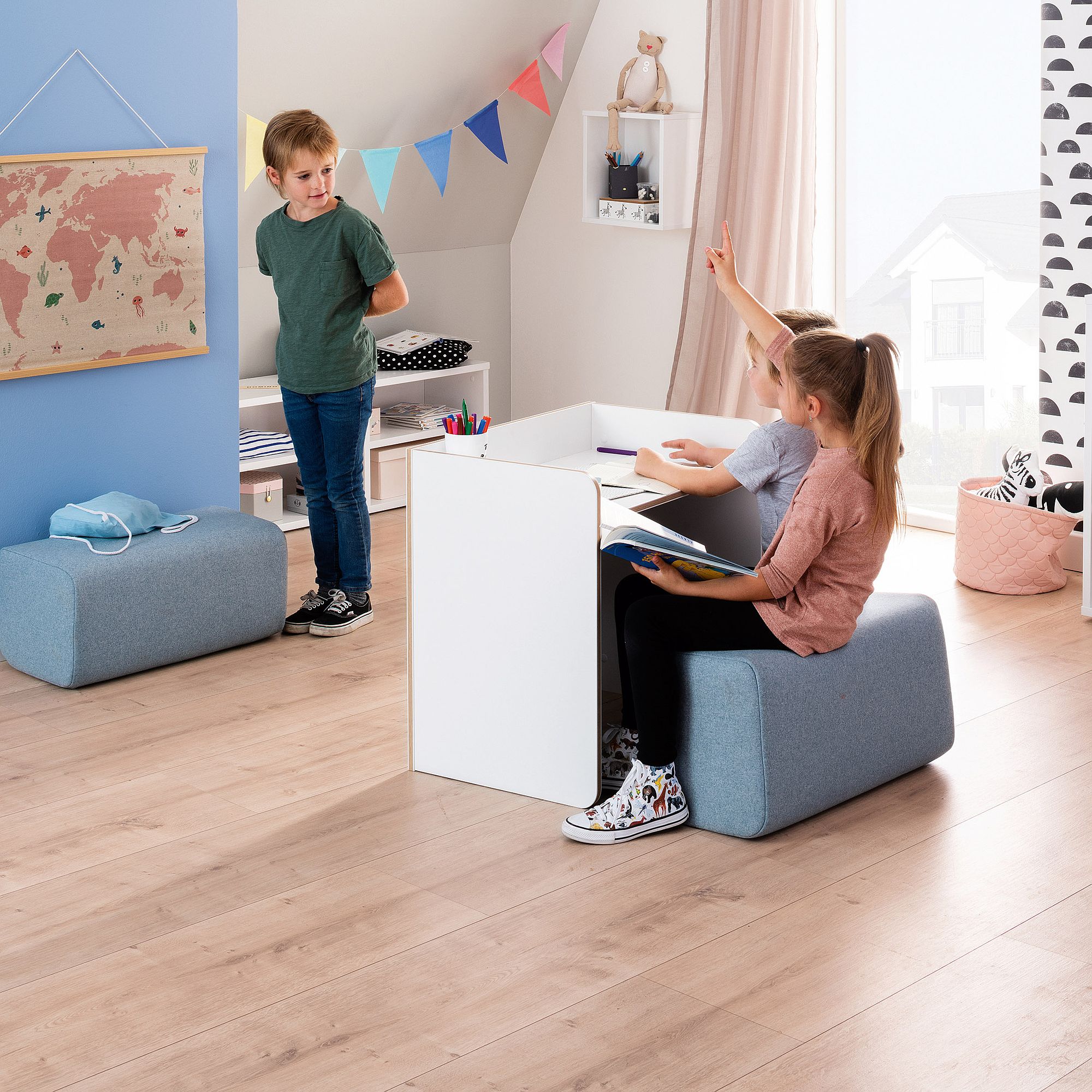 Zwei Kinder sitzen an einem kleinen Schreibtisch, während ein anderes Kind neben einer Wandkarte steht.