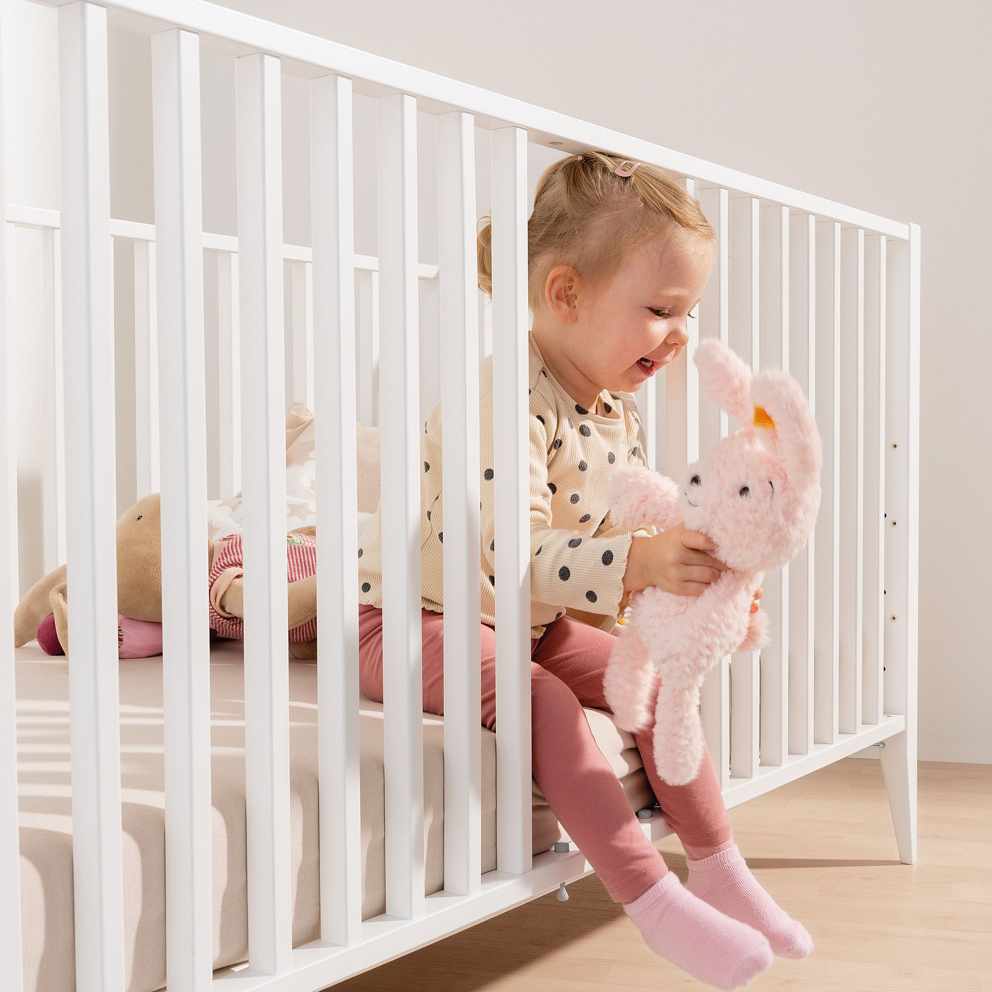Babybett Felie 70 x 140 cm Kleinkind sitzt auf dem Rand des Kinderbettes, hält einen rosa Stoffhasen und lächelt.