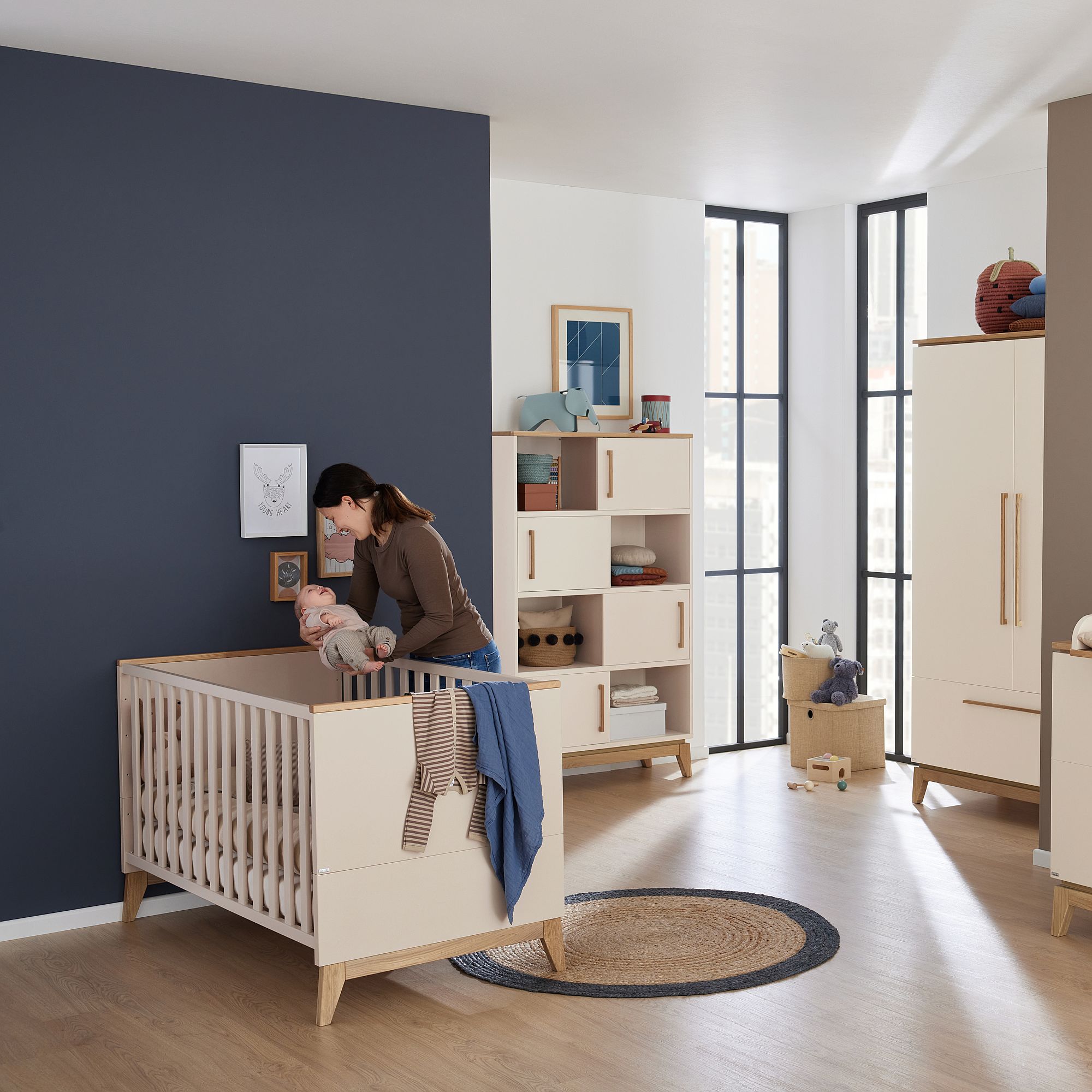 Eine Frau kümmert sich in einem modernen Kinderzimmer mit hellen Holzmöbeln um ihr Baby.