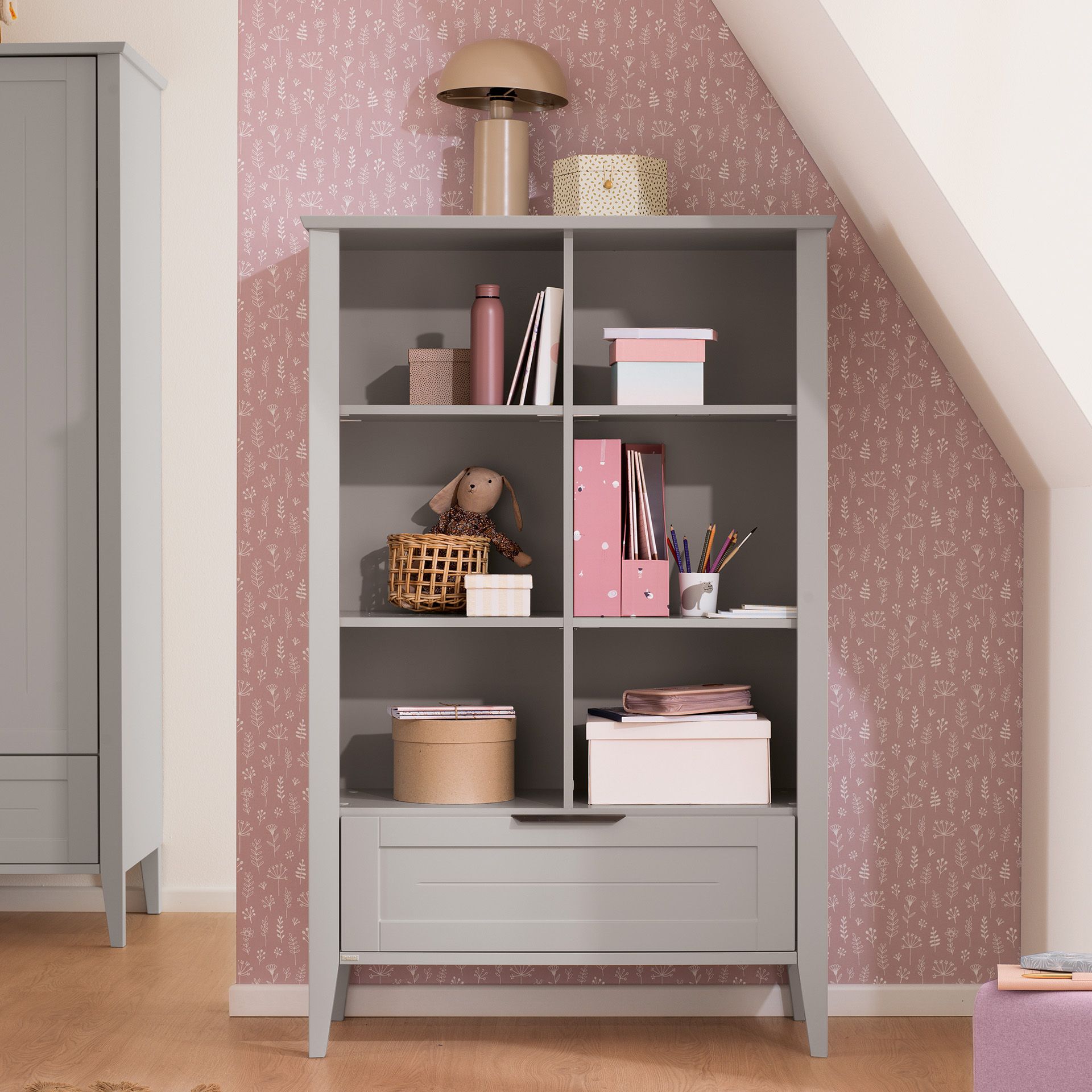 Gray bookshelf with boxes, a lamp, toy, and decor against pink patterned wall.