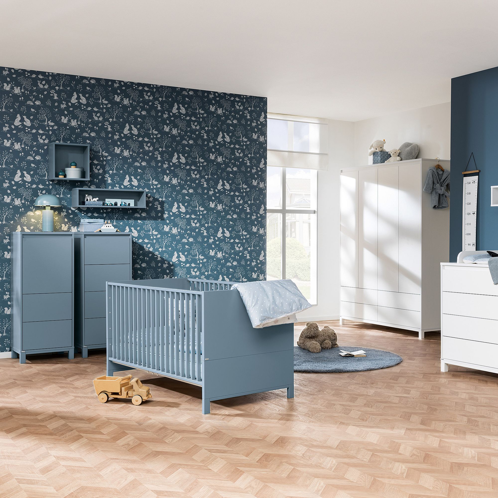 Blau-weißes Kinderzimmer mit Kinderbett, Kommoden, Spielzeug und gemusterter Tapete.
