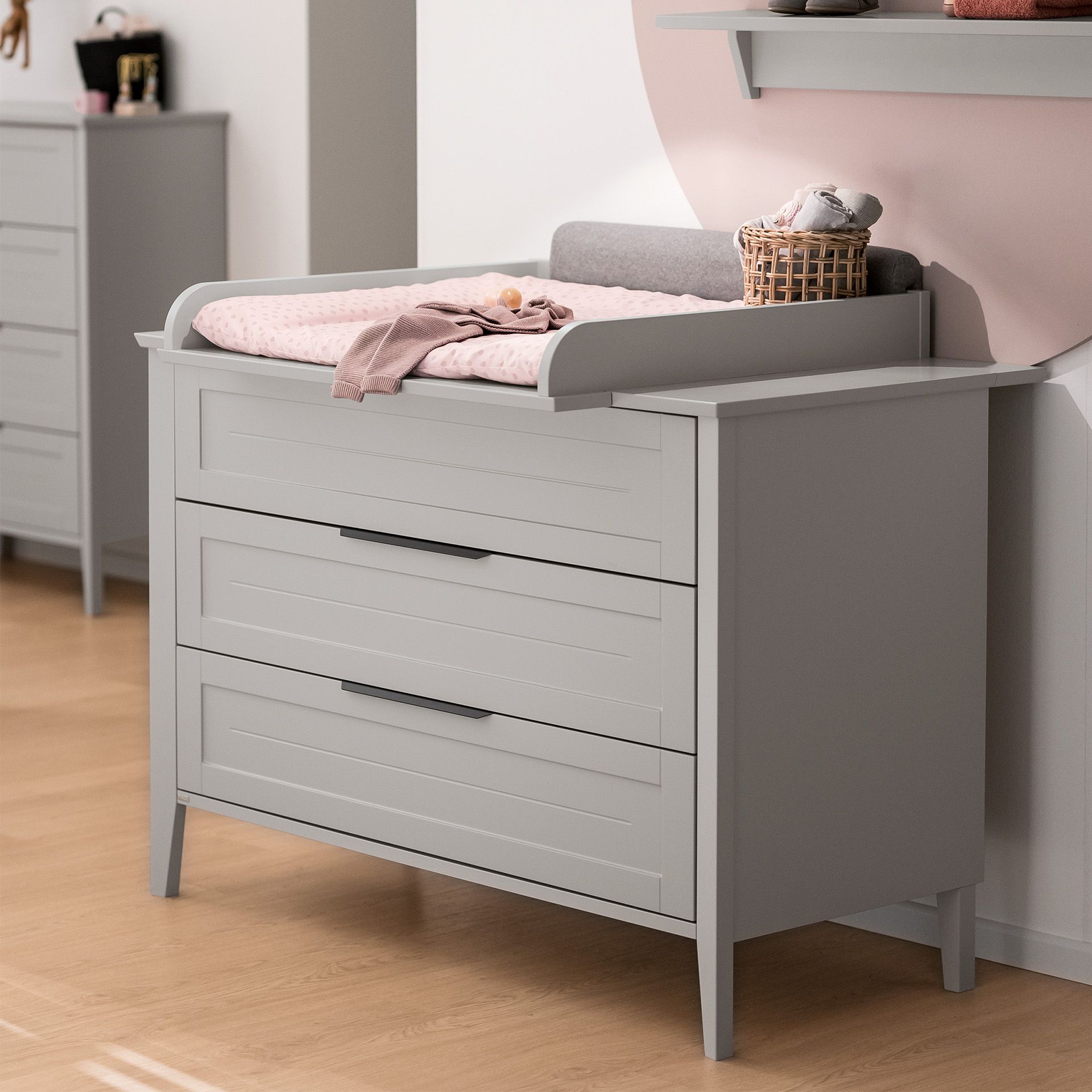 Wickelaufsatz Levke zur Erweiterung der Wickelkommode mit 3 Schubladen Grauer Wickeltisch mit einer rosafarbenen Matte und einem kleinen Korb darauf in einem Kinderzimmer.