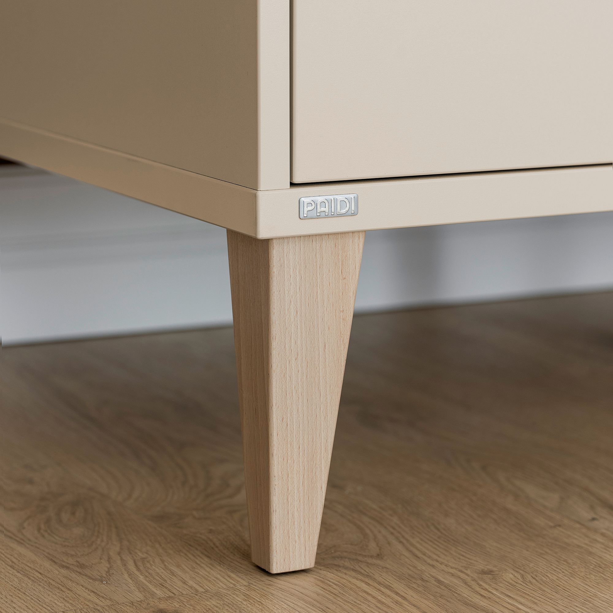 Beiger Schrank mit Holzbeinen auf einem Holzboden, PAIDI-Etikett an der Vorderseite.