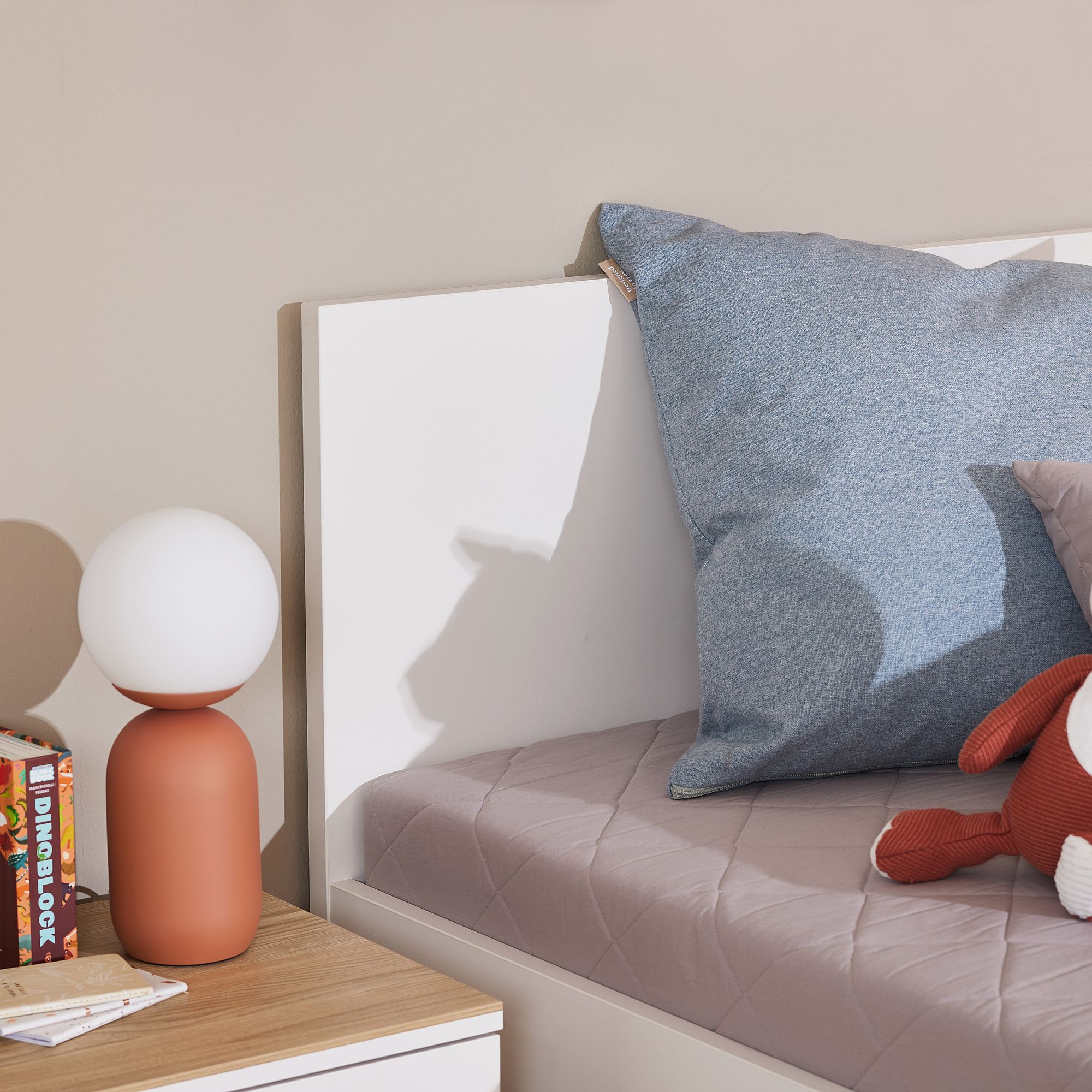 Ein gemütliches Bett mit blauen Kissen, eine rote Lampe, Bücher und ein Spielzeug auf einem Nachttisch.