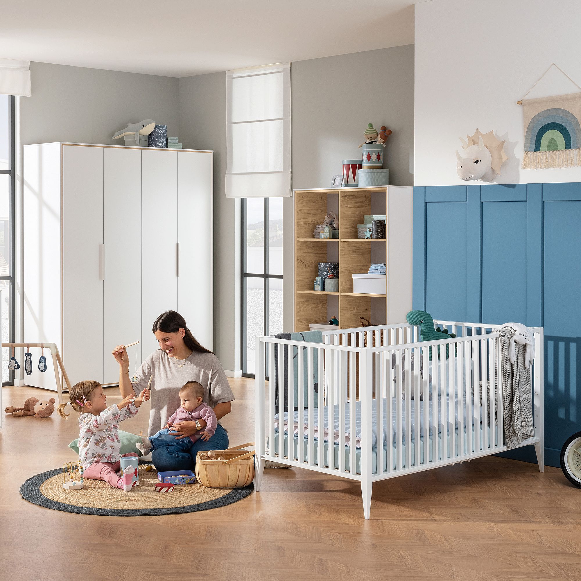 Eine Mutter sitzt mit zwei Kindern auf dem Boden neben einem Kinderbett in einem Kinderzimmer.