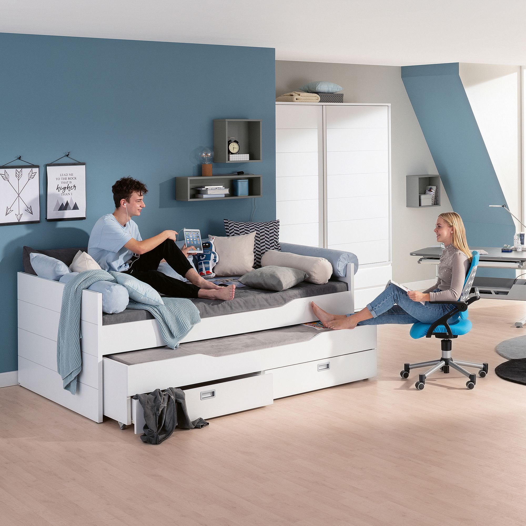 Ein Teenager sitzt auf einem weißen Tagesbett und unterhält sich mit einem Mädchen, das auf einem blauen Schreibtischstuhl sitzt.
