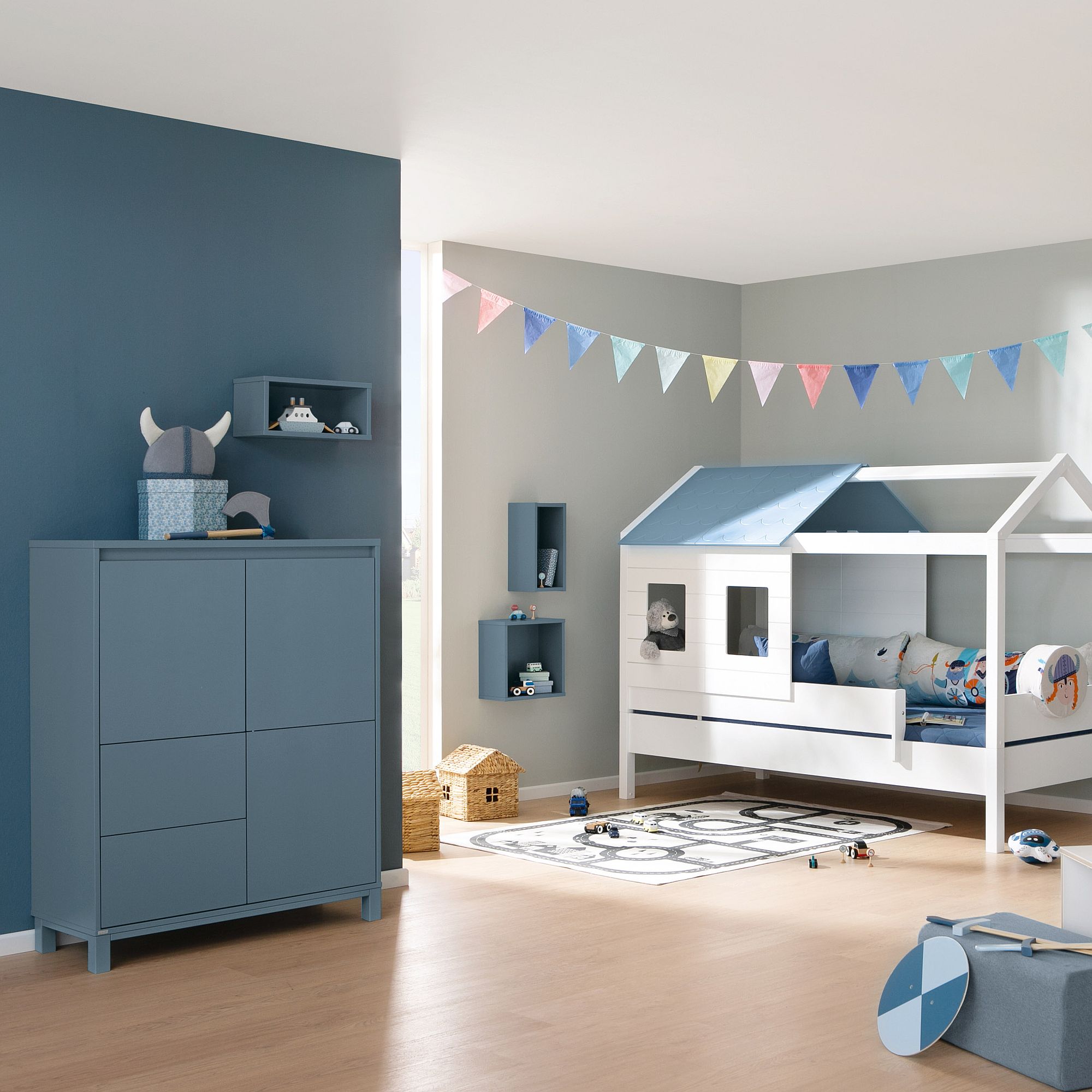 Kinderzimmer mit blauer Einrichtung, einem Bett in Hausform, Spielzeug und bunten Wimpelketten.
