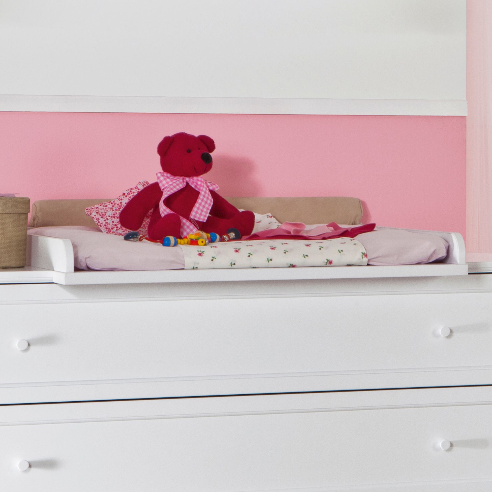 Red teddy bear and toys on a white dresser with a pink wall in the background.
