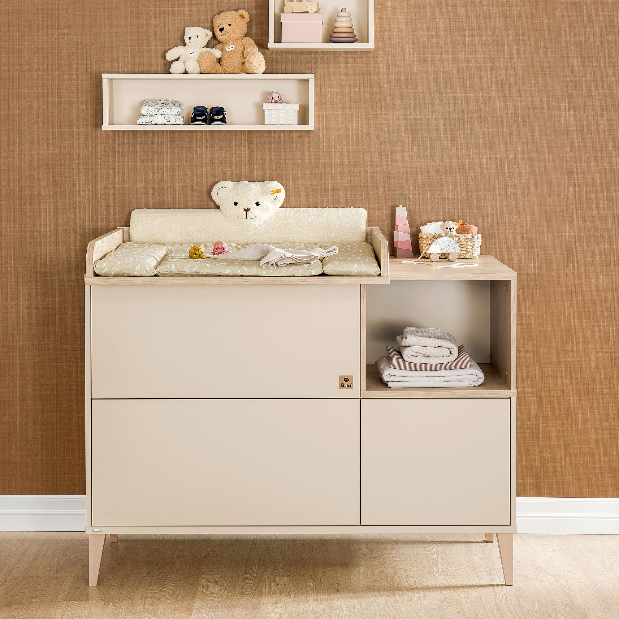 Beigefarbener Wickeltisch mit Teddybären, Regalen und gefalteter Kleidung.