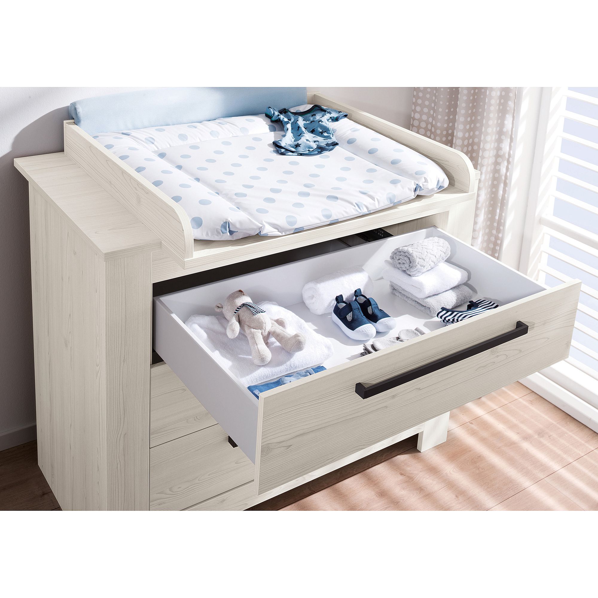 Weißer Wickeltisch mit einer offenen Schublade für Babykleidung und ein Stofftier.