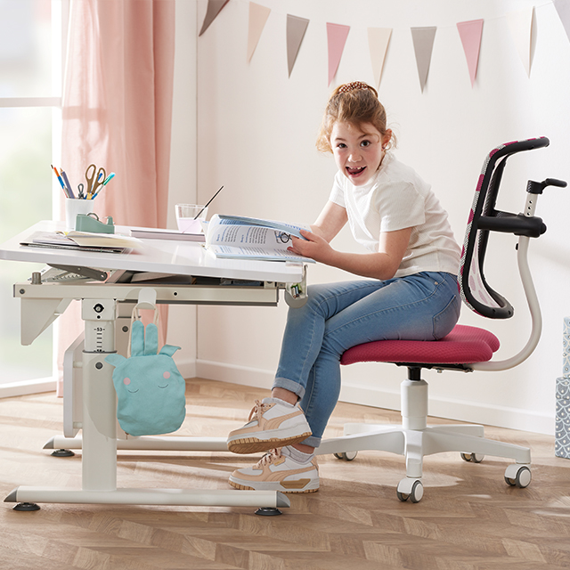Smiling girl sitting at a desk reading a book in a decorated room.