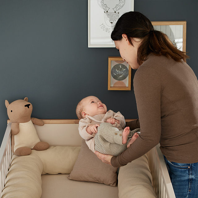 1106_PAIDI_Shop_Start_Feed_Babybetten-ID1697554_120d4e84-5022-3dba-1a19-a90fa21c12c0.jpg Frau beugt sich über den Wickeltisch und hält ein Baby, in der Nähe ein Stofftier.