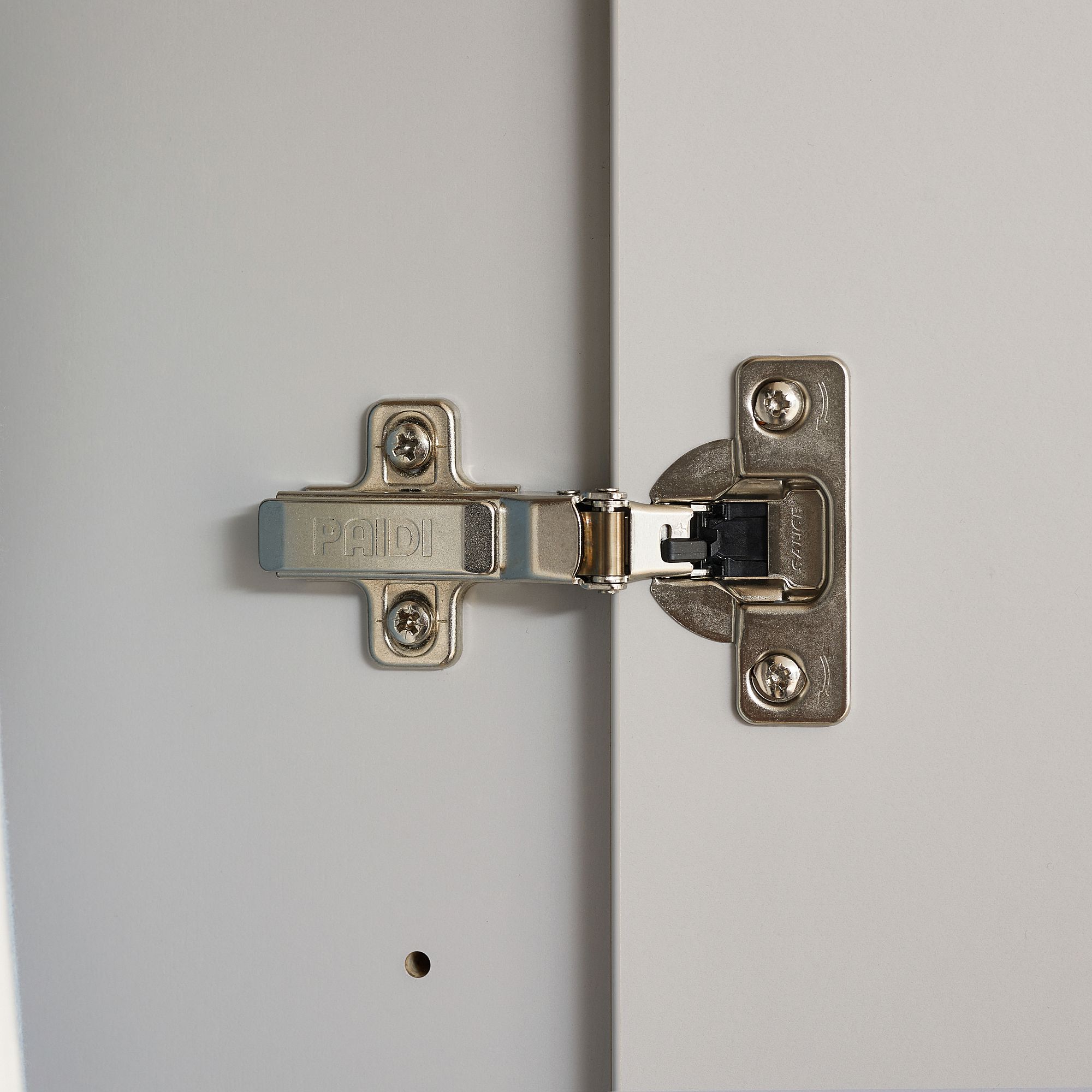 Drehtürenschrank Fiene mit 4 Türen und 1 Schublade Nahaufnahme eines Metallscharniers, das an zwei weißen Platten befestigt ist.