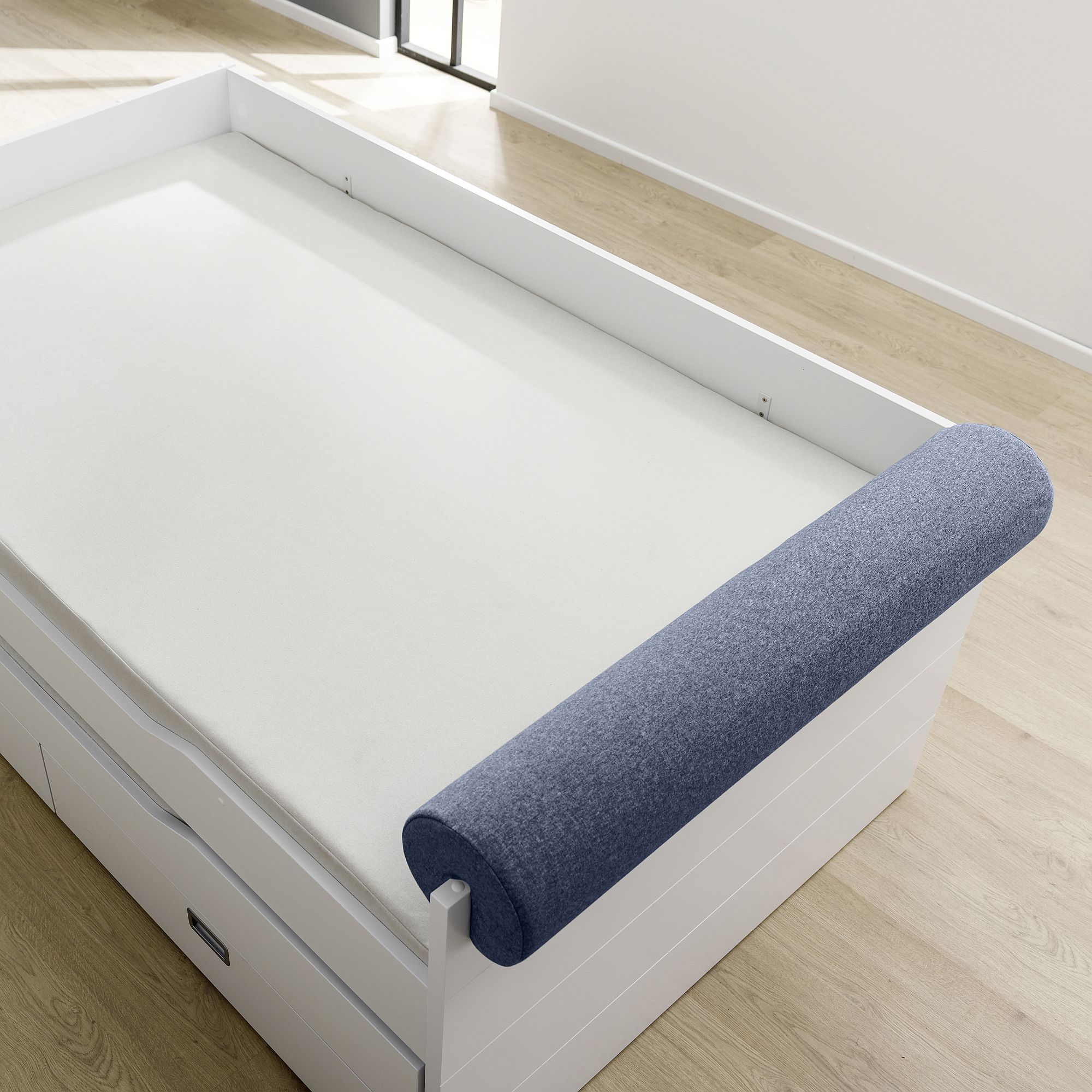 White bed with a rolled blue cushion on one side, in a bright room with wood floor.