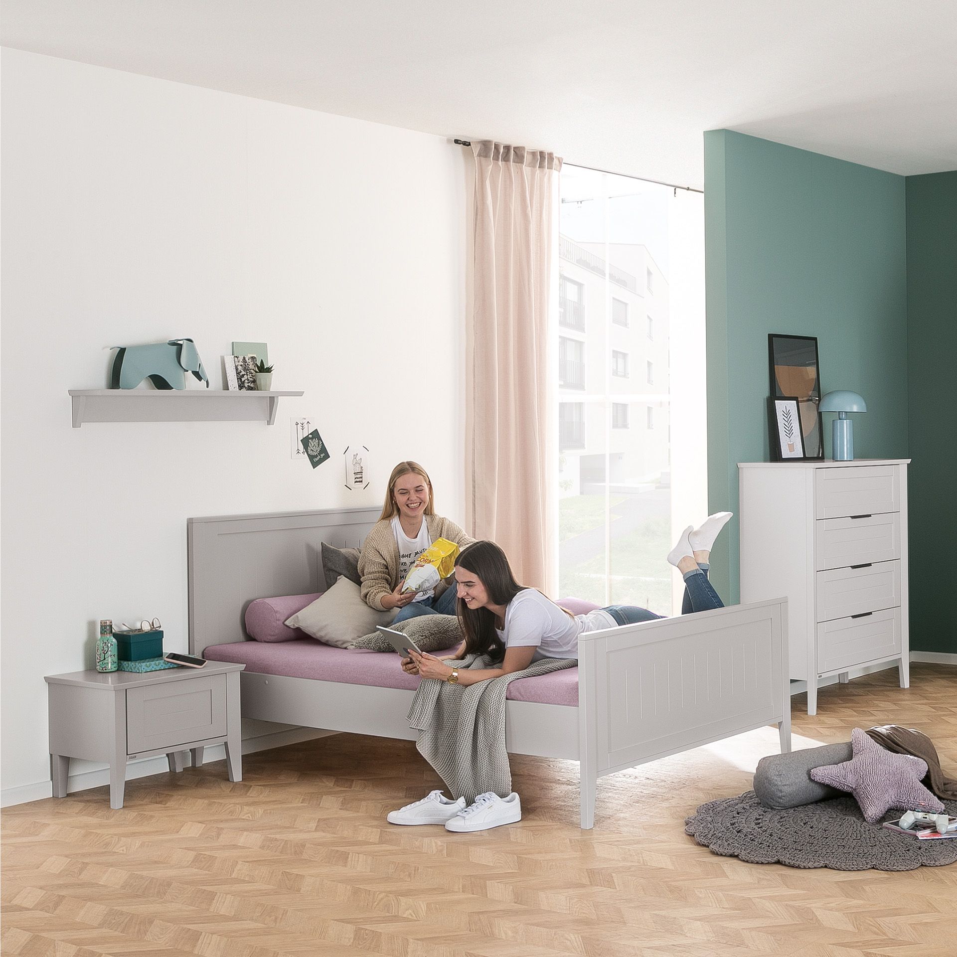 Two young women relax on a bed in a bright, modern bedroom with light furniture.