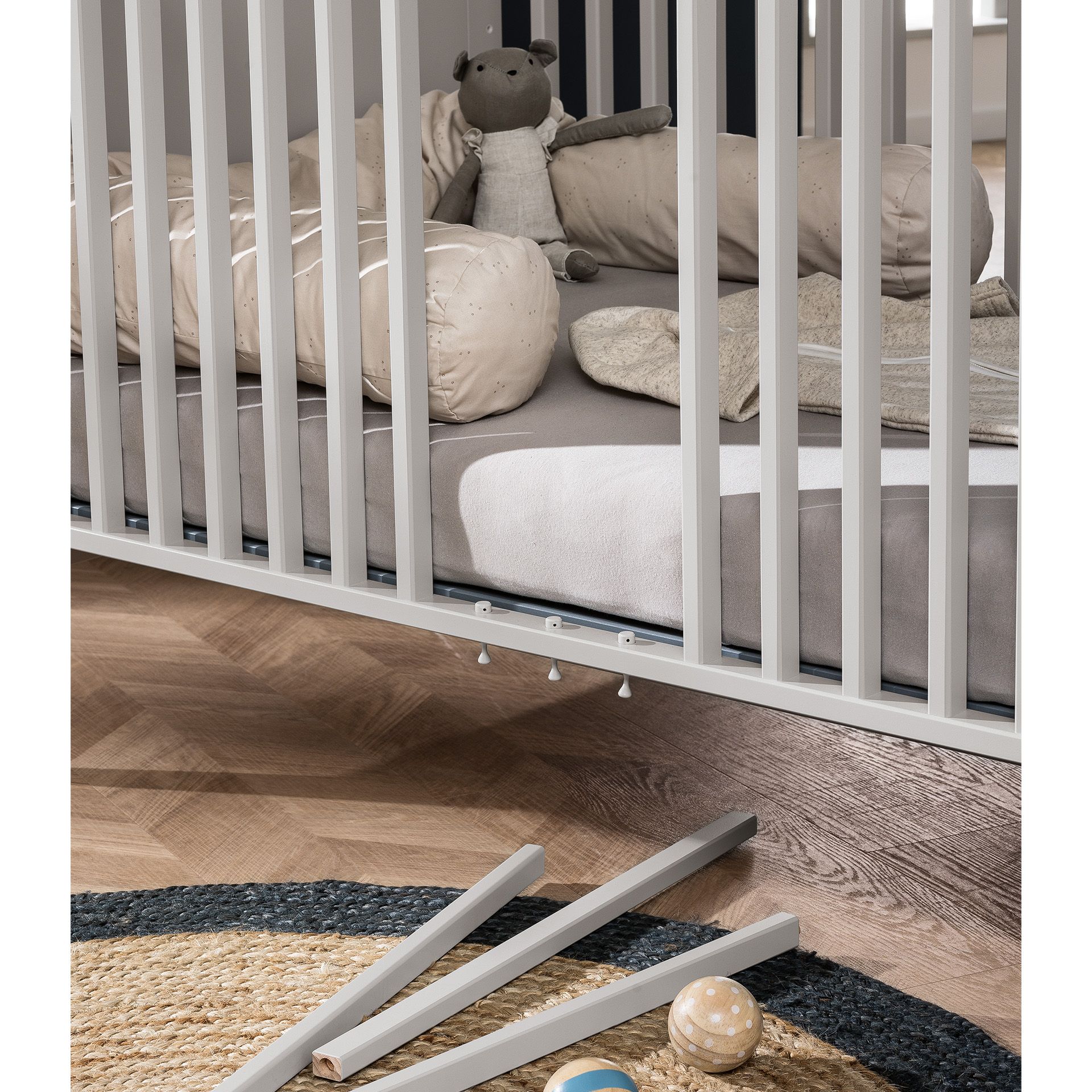 Babybett Levke 70 x 140 cm Graues Kinderbett mit fehlenden Latten, Plüschtier, Bettzeug und Spielzeug auf dem Boden.