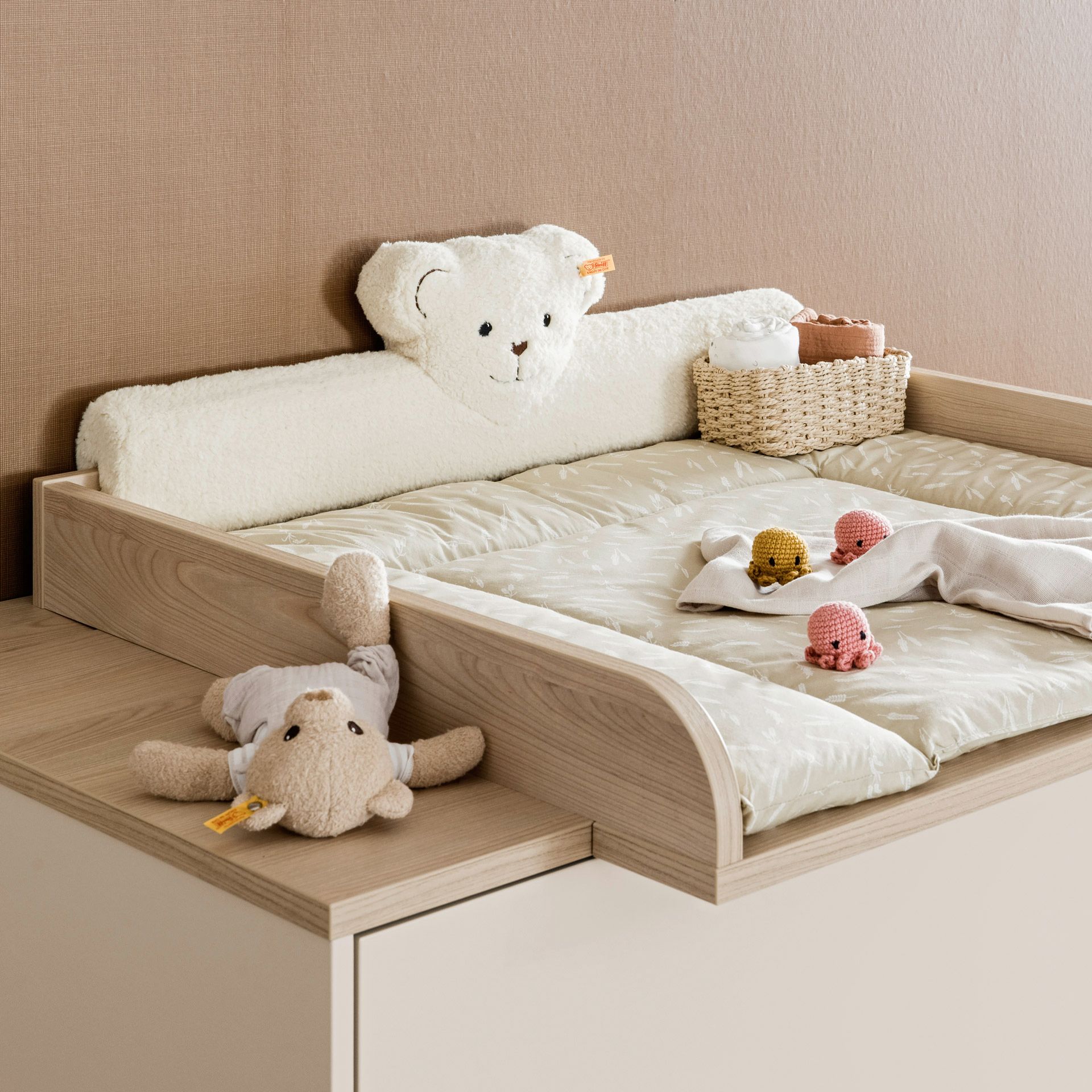 Kopfschutz Steiff by PAIDI Kissen für Wickelaufsatz, Beige A baby changing table with a bear pillow, toys, and a basket on top.
