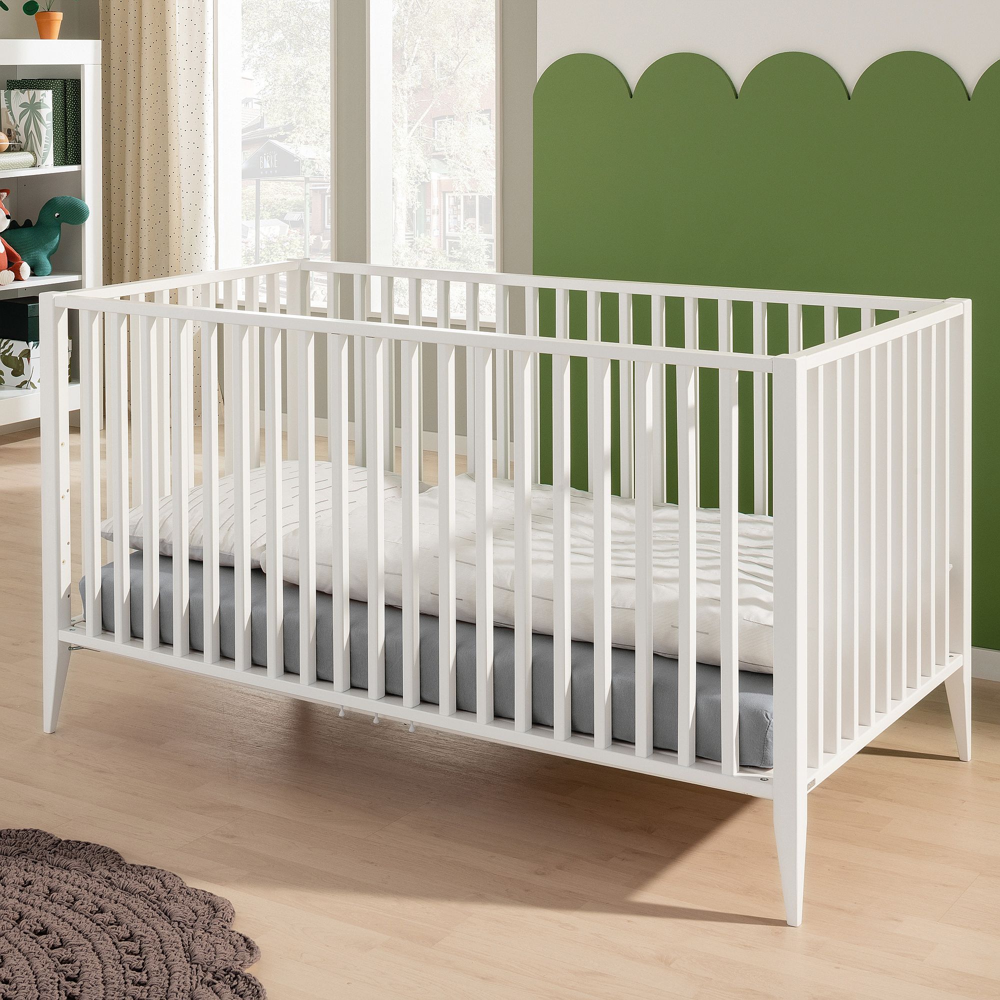Weißes Babybett mit gestreifter Bettwäsche in einem Kinderzimmer mit grünen und weißen Wänden.