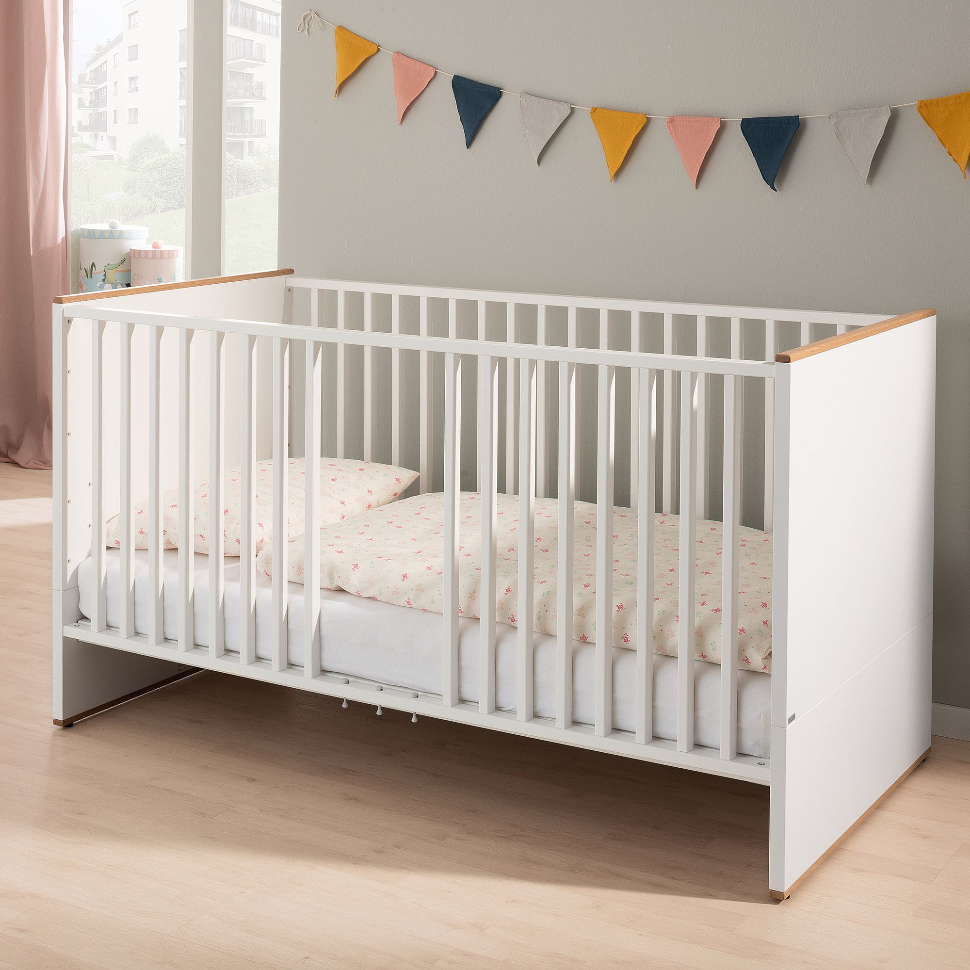 Weißes Babybett mit Bettwäsche, neben einer Wand, die mit bunten Wimpeln geschmückt ist.