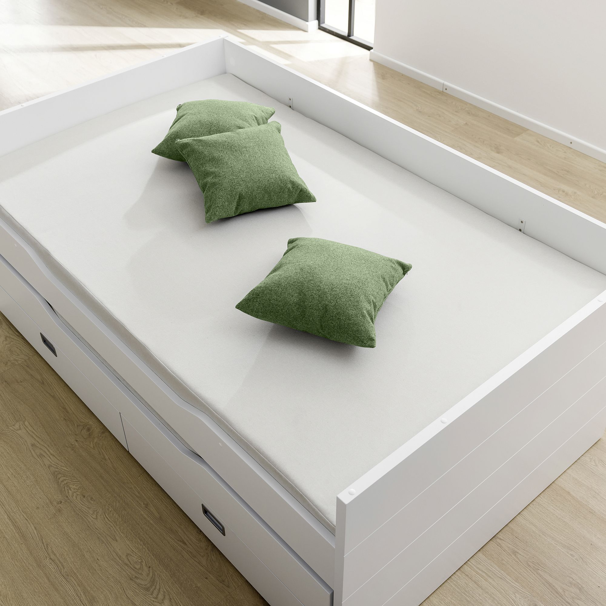 White bed with three green pillows in a bright room with wood flooring.
