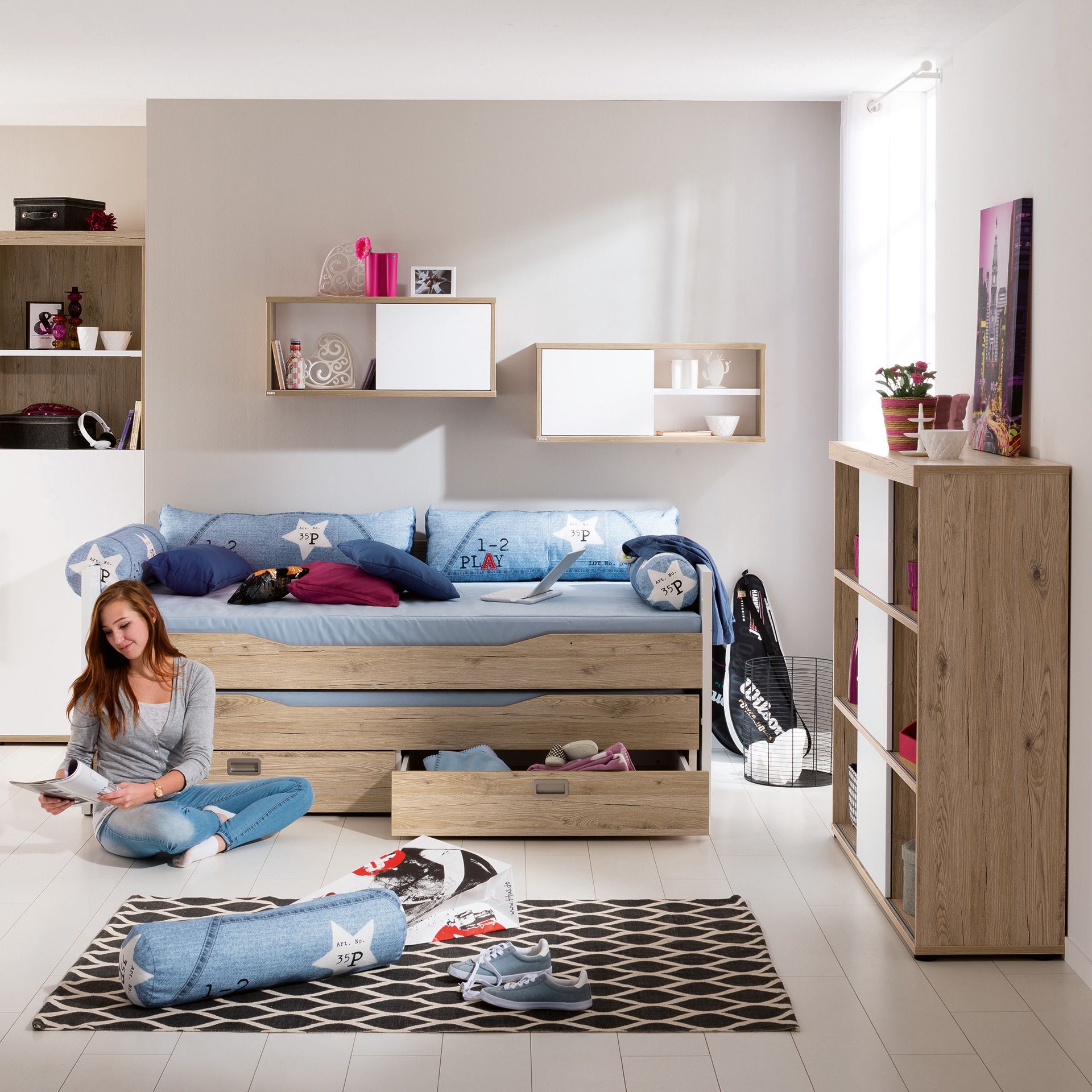Eine junge Frau liest auf dem Boden in einem modernen, gemütlichen Schlafzimmer mit blauer und holzfarbener Einrichtung.