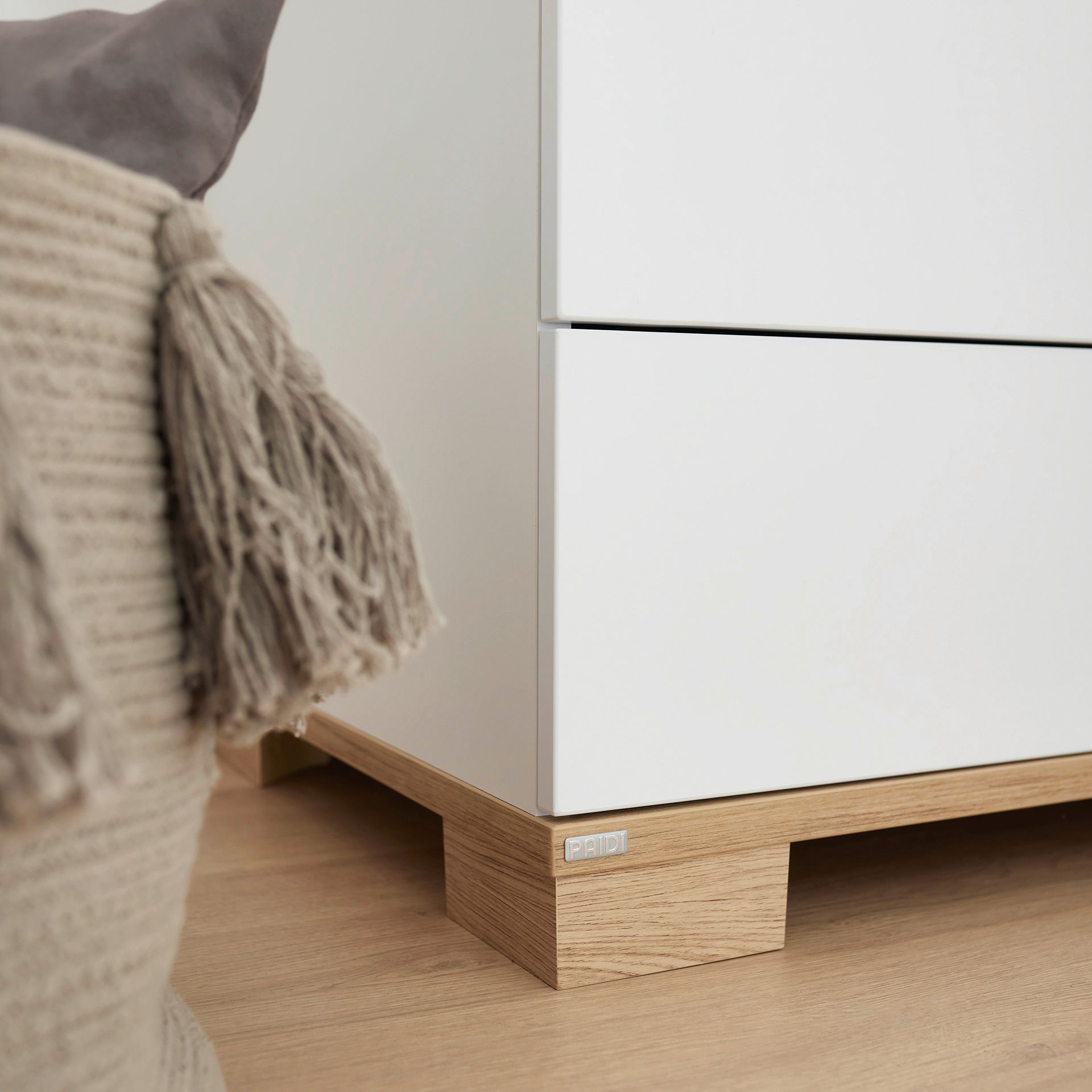 Close-up of a white drawer unit with wooden base next to a tasseled woven basket.