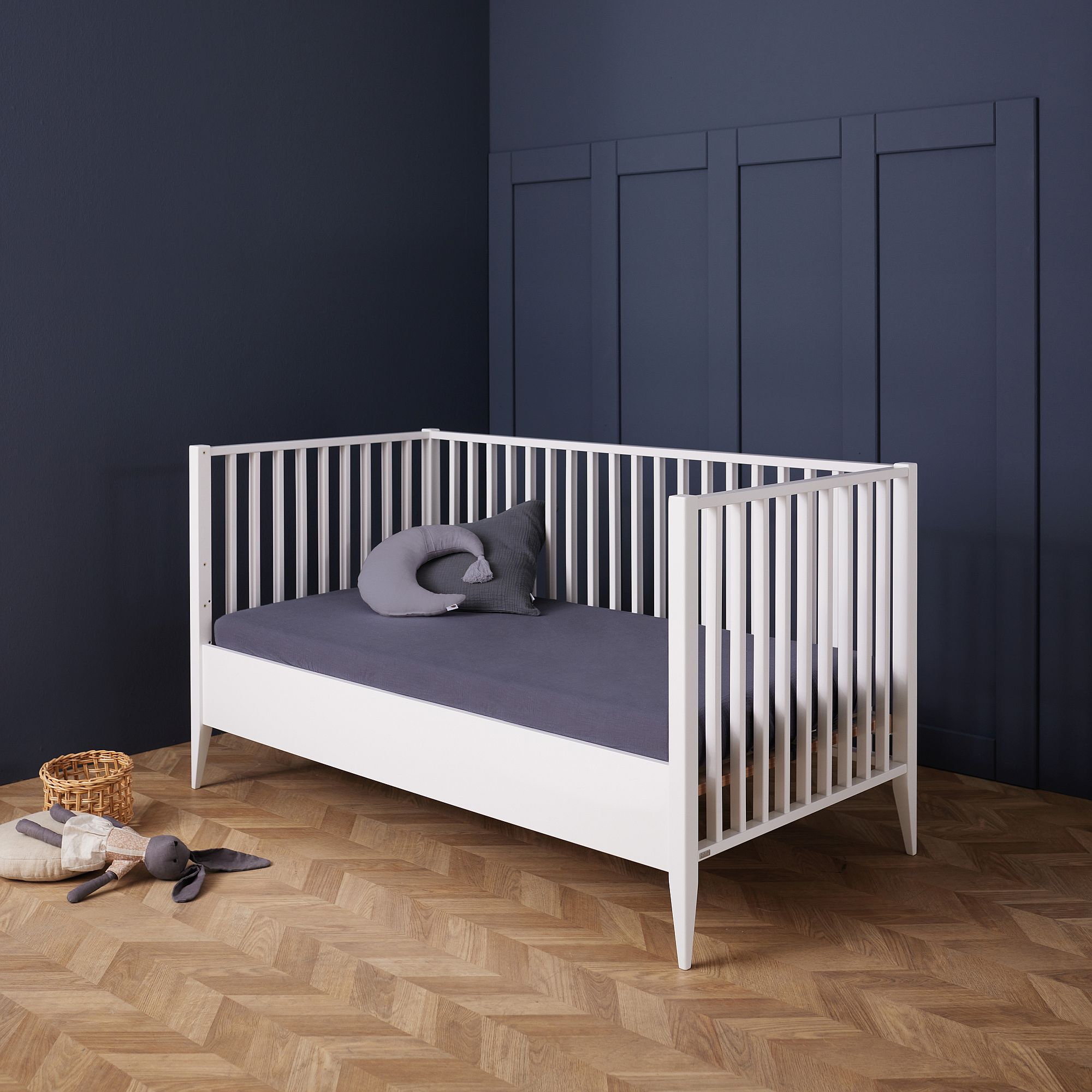 Weißes Kinderbett mit dunkler Bettwäsche und Kissen in einem navyblauen Zimmer mit Holzboden.