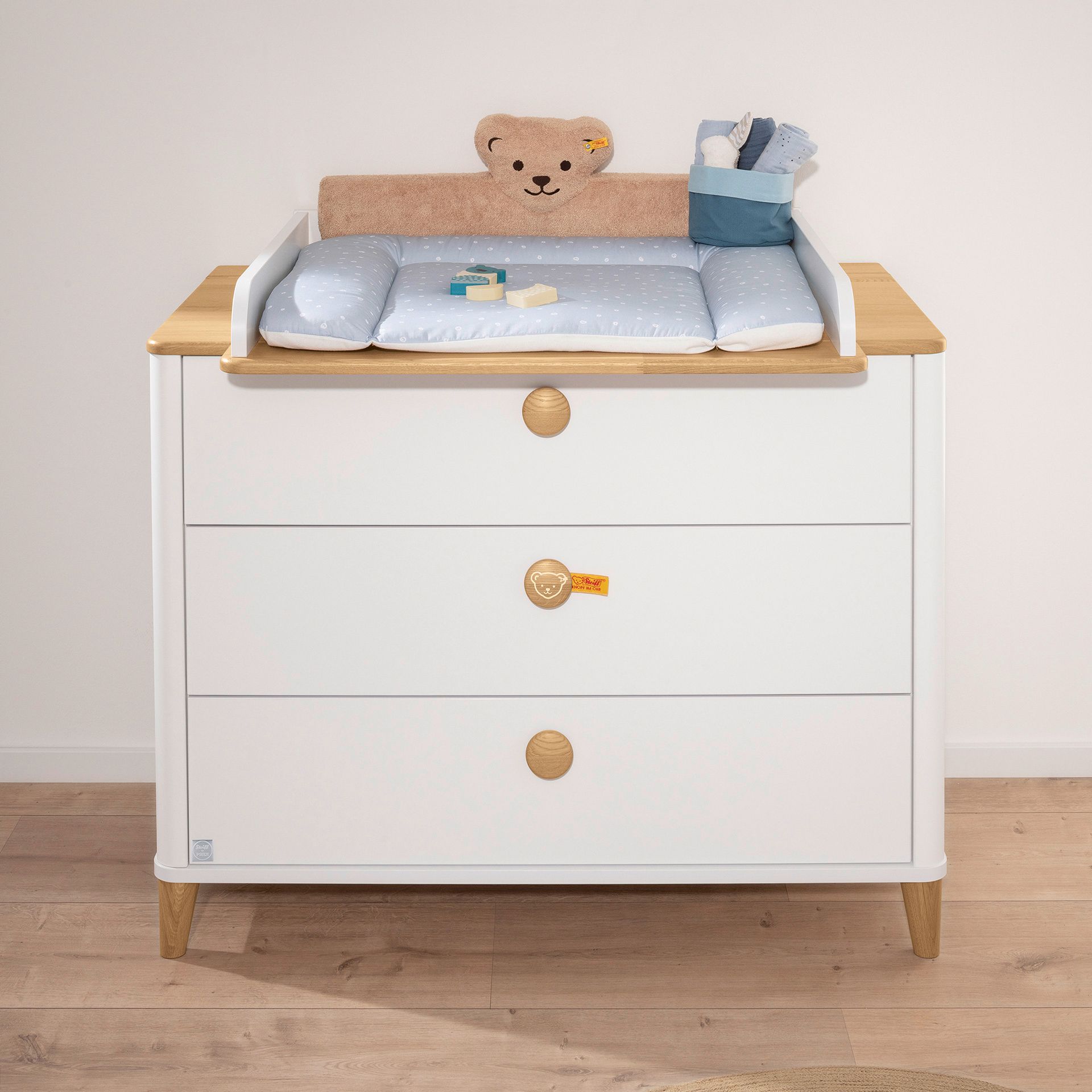 Wickelaufsatz Lotte & Fynn - Steiff by PAIDI zur Erweiterung der Wickelkommode mit 3 Schubladen Weißer Wickeltisch mit einer blauen Matte, einem Teddybär-Kissen und Babyutensilien darauf.