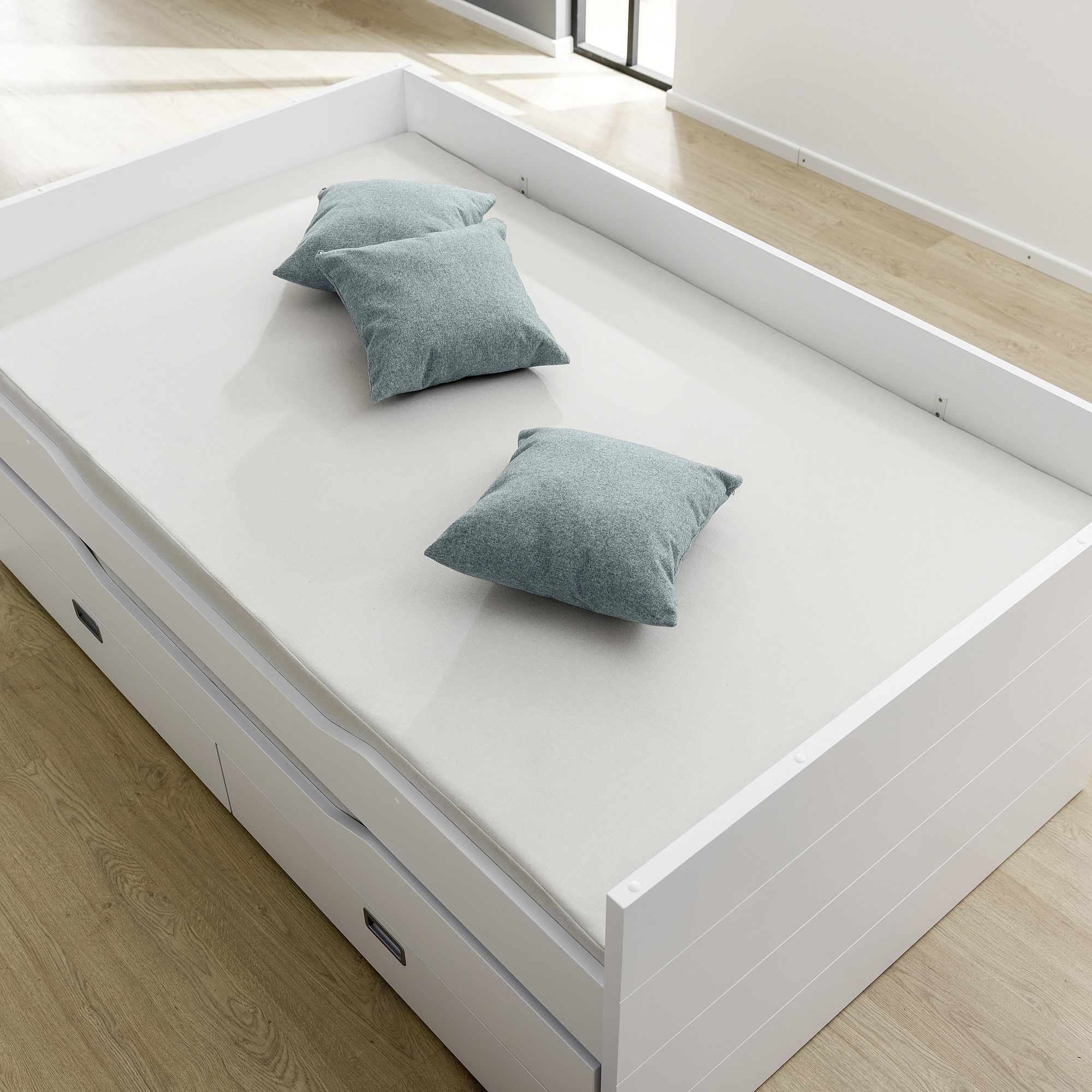 White daybed with three gray pillows on a light wood floor in a bright room.