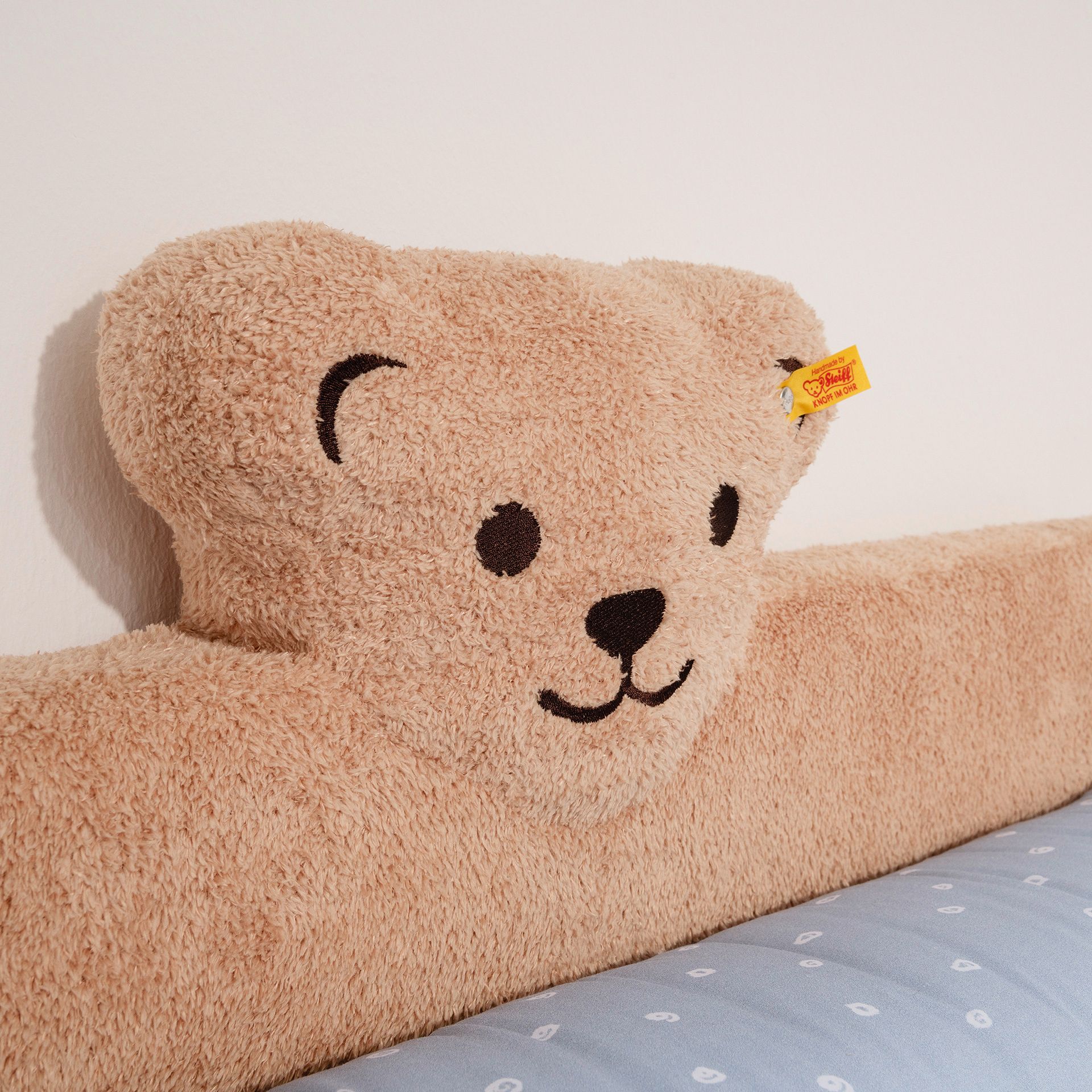 Kopfschutz  als Kantenschutz für den Wickelaufsatz mit Steiff-Plüsch-Teddy in Braun Plüschkissen in Form eines Teddybären, das an einer weißen Wand über einem blauen Bett ruht.