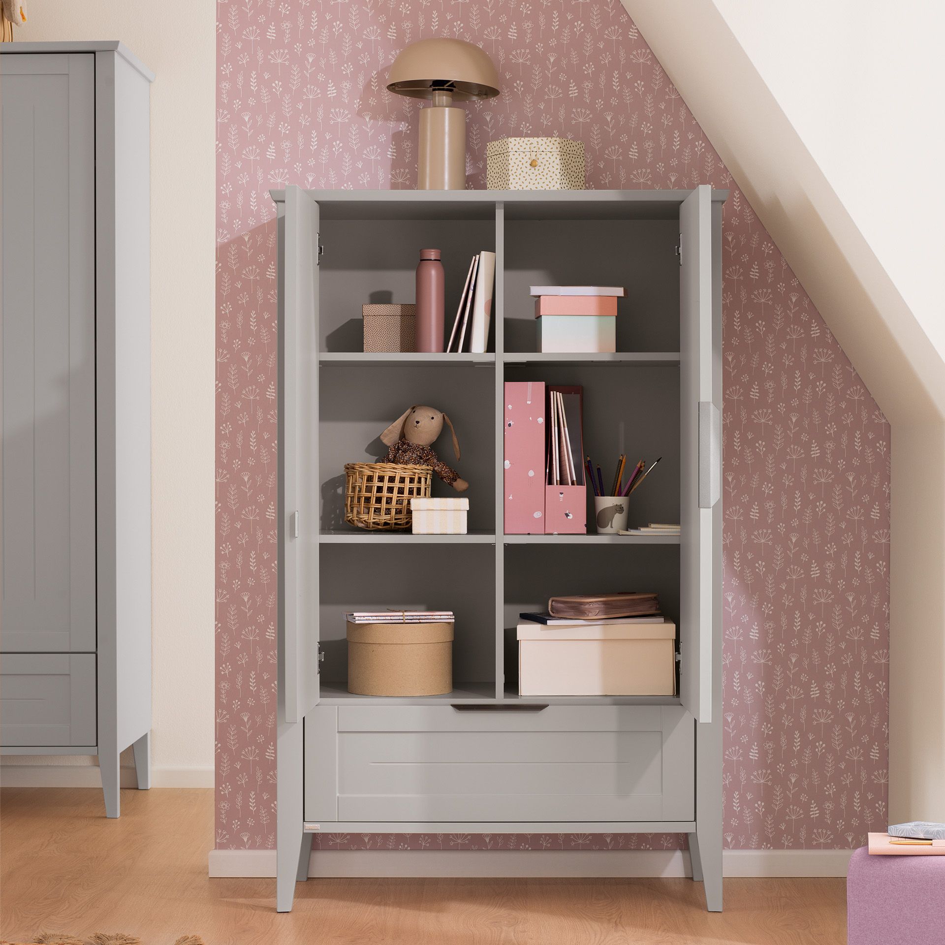 Drehtürenschrank Levke in der Tiny-Größe mit 2 Türen und 1 Schublade Grauer Schrank mit Regalen mit Büchern, Kisten, einer Lampe und einem ausgestopften Hasen.