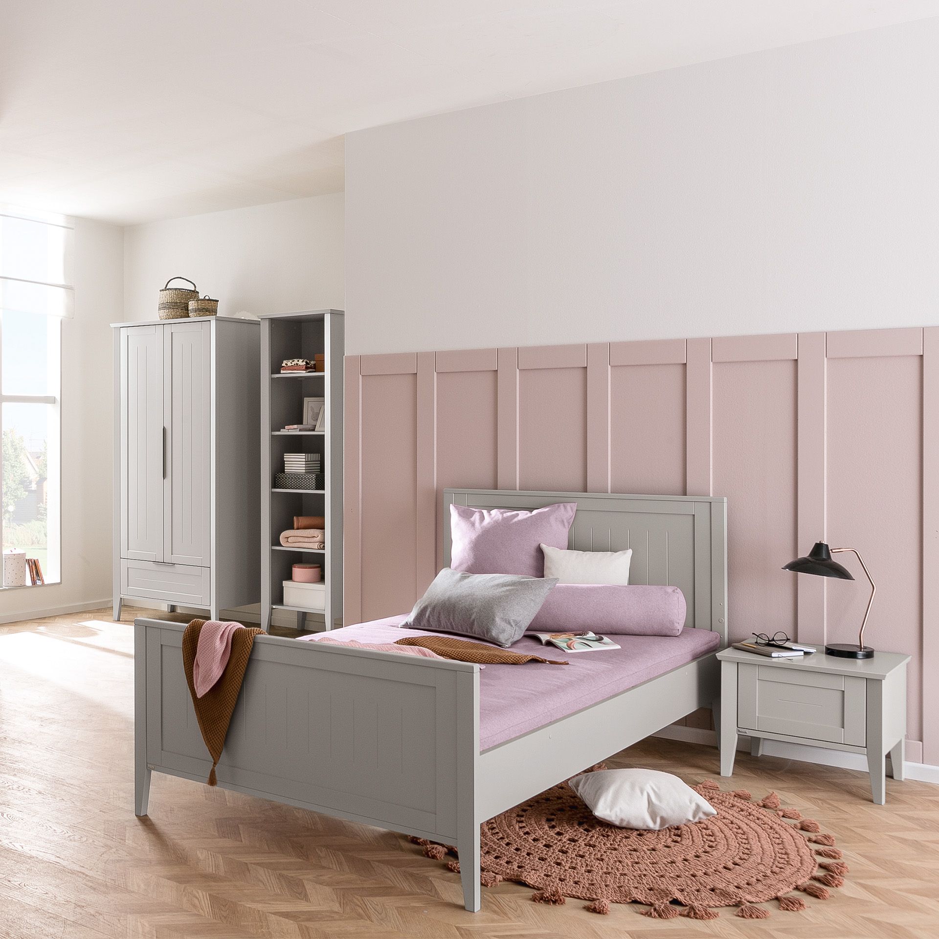 Bett Levke Kinderbett in 140 x 200 cm Ein gemütliches Schlafzimmer mit grauem Bett, rosafarbener Bettwäsche, Teppich und Kleiderschrank in sanfter Beleuchtung.
