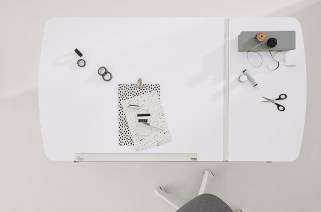 Minimalist white desk with scattered papers, tape, scissors, a pen, and a small gray organizer, viewed from above.