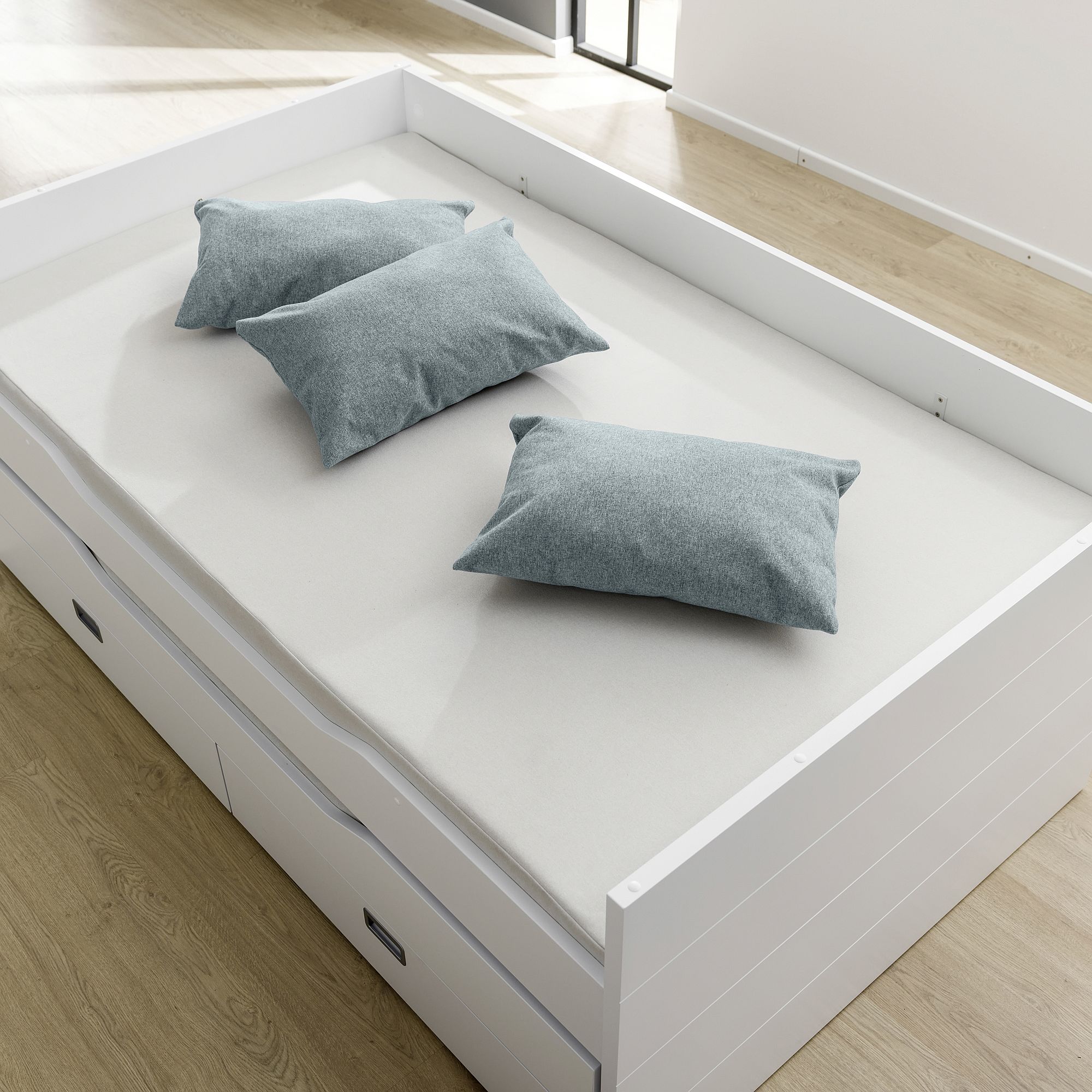 A white bed with three gray pillows on a light wood floor.