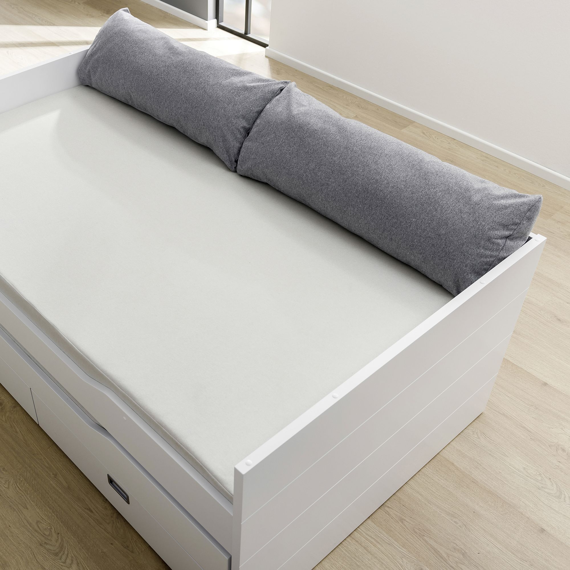 White daybed with storage drawers, light mattress, and a long gray pillow.