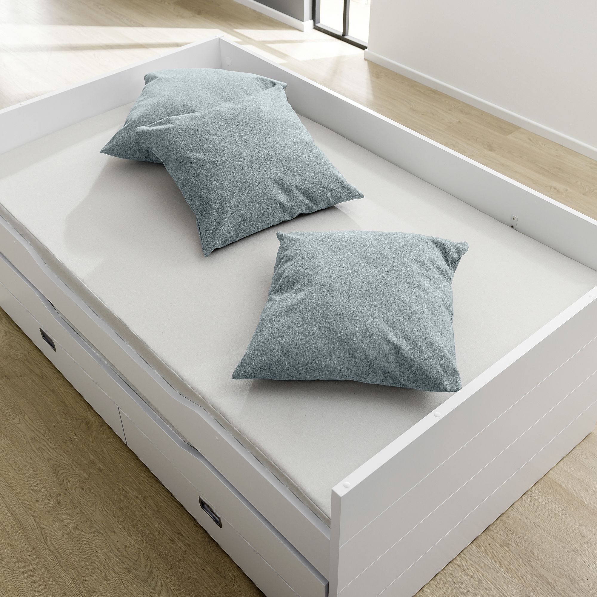 White bed with two gray pillows on a light wooden floor in a bright room.