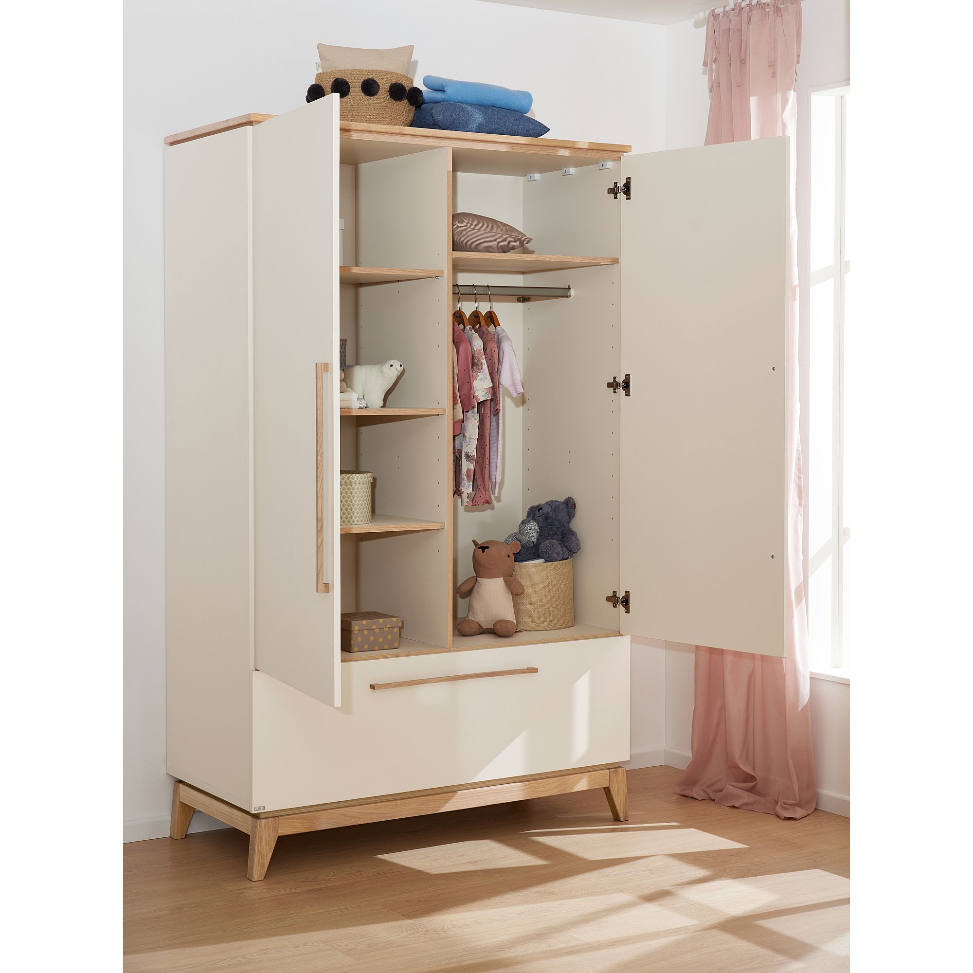 Offener Kleiderschrank mit Regalen, in dem Babykleidung, Spielzeug und gefaltete Decken aufgehängt sind.