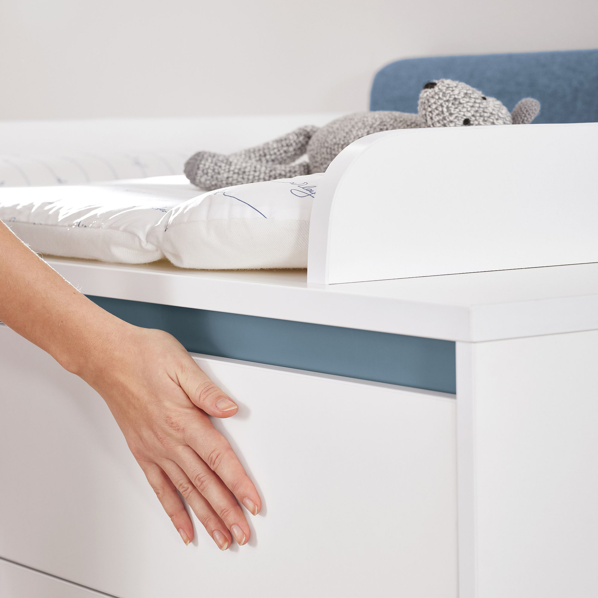 Eine Hand öffnet eine weiße Kommodenschublade neben einem Wickeltisch mit einem Stofftier.