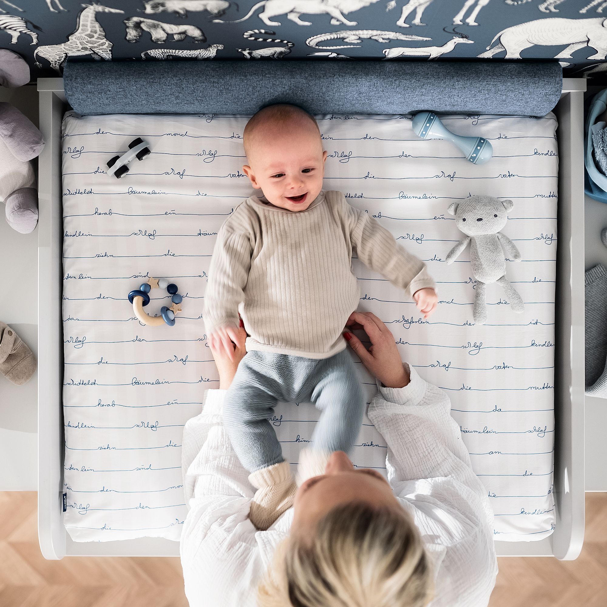 A smiling baby lies on a changing table, held by an adult, with toys nearby.