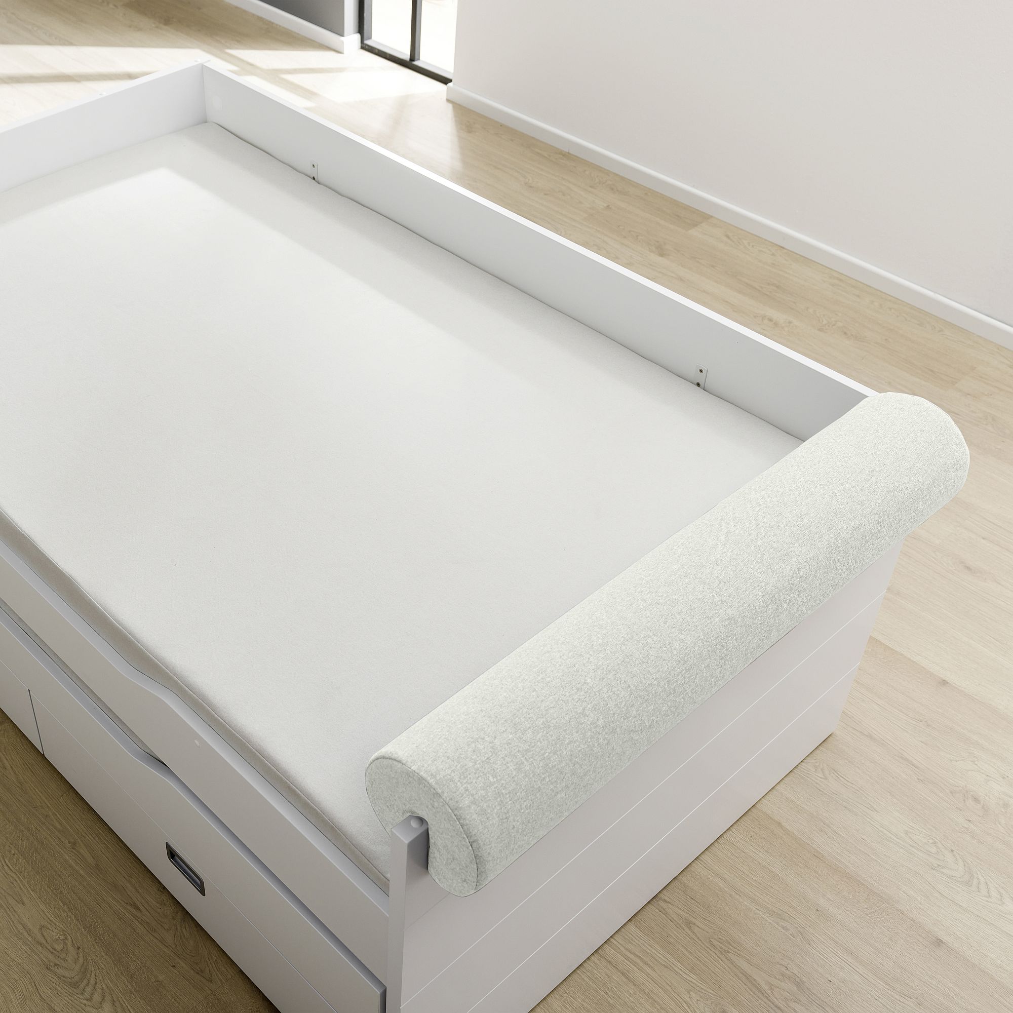 A rolled-up mattress topper on a white bed with drawers in a bright room.