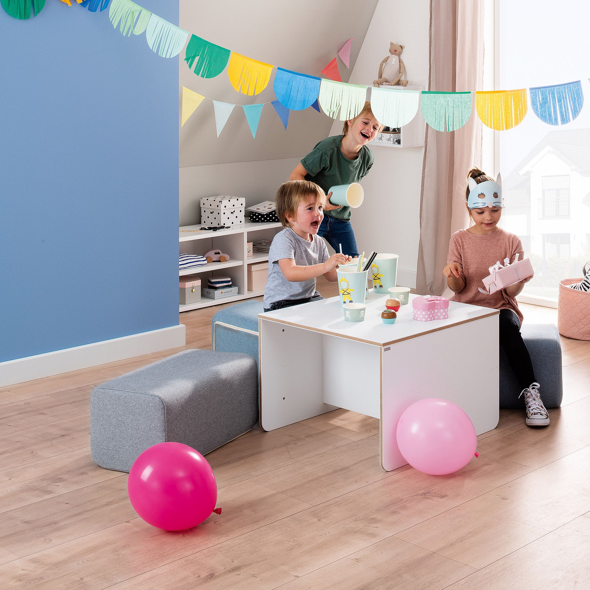 Drei Kinder spielen an einem Tisch mit Luftballons und Dekorationen in einem hellen Raum.